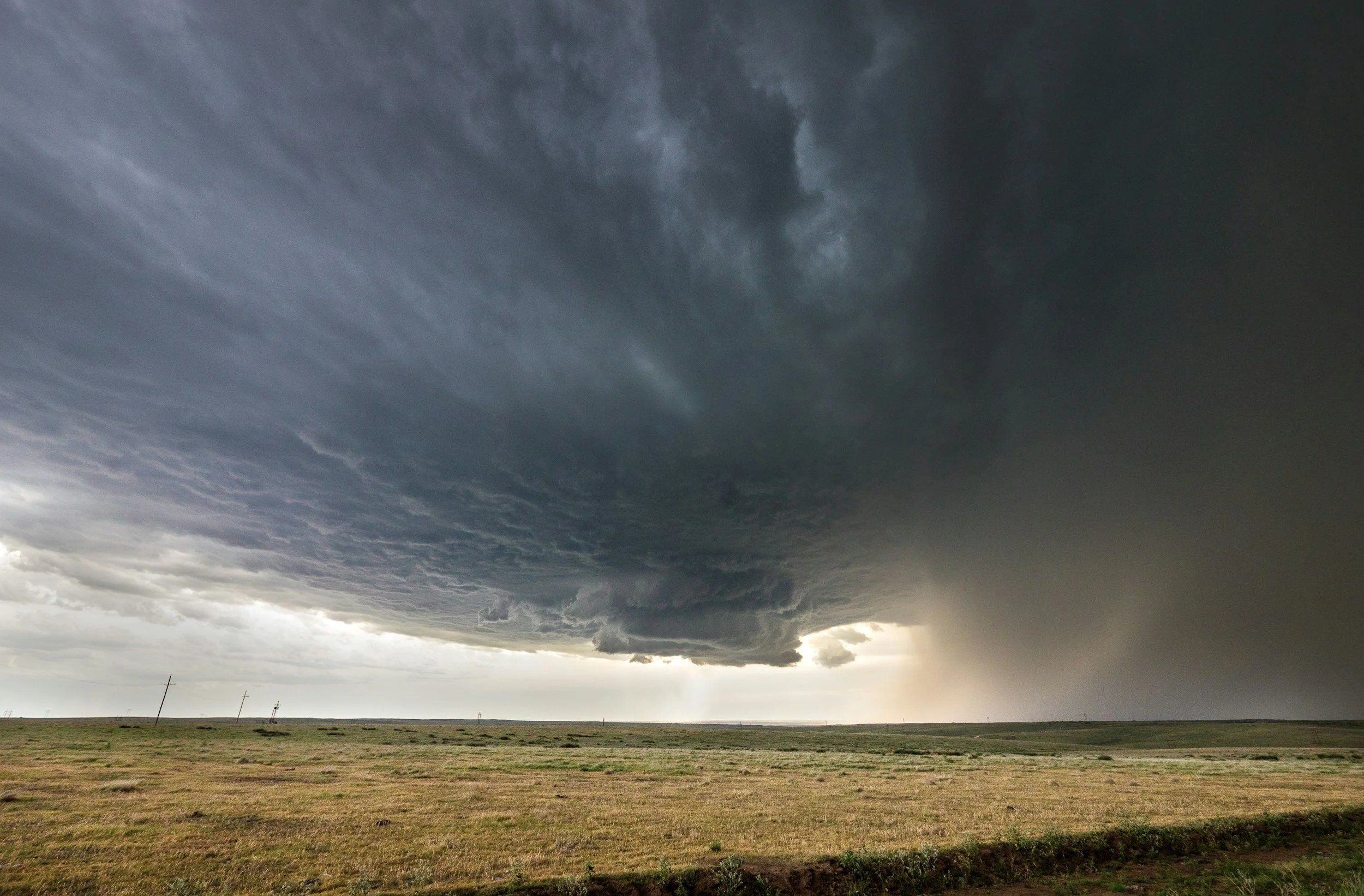 Dramatic dark storm clouds over a flat grassy plain with distant power poles and sunlight breaking through the horizon