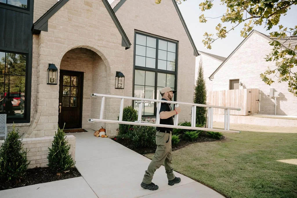 Man carrying an extension ladder past the entrance of a modern stone house.