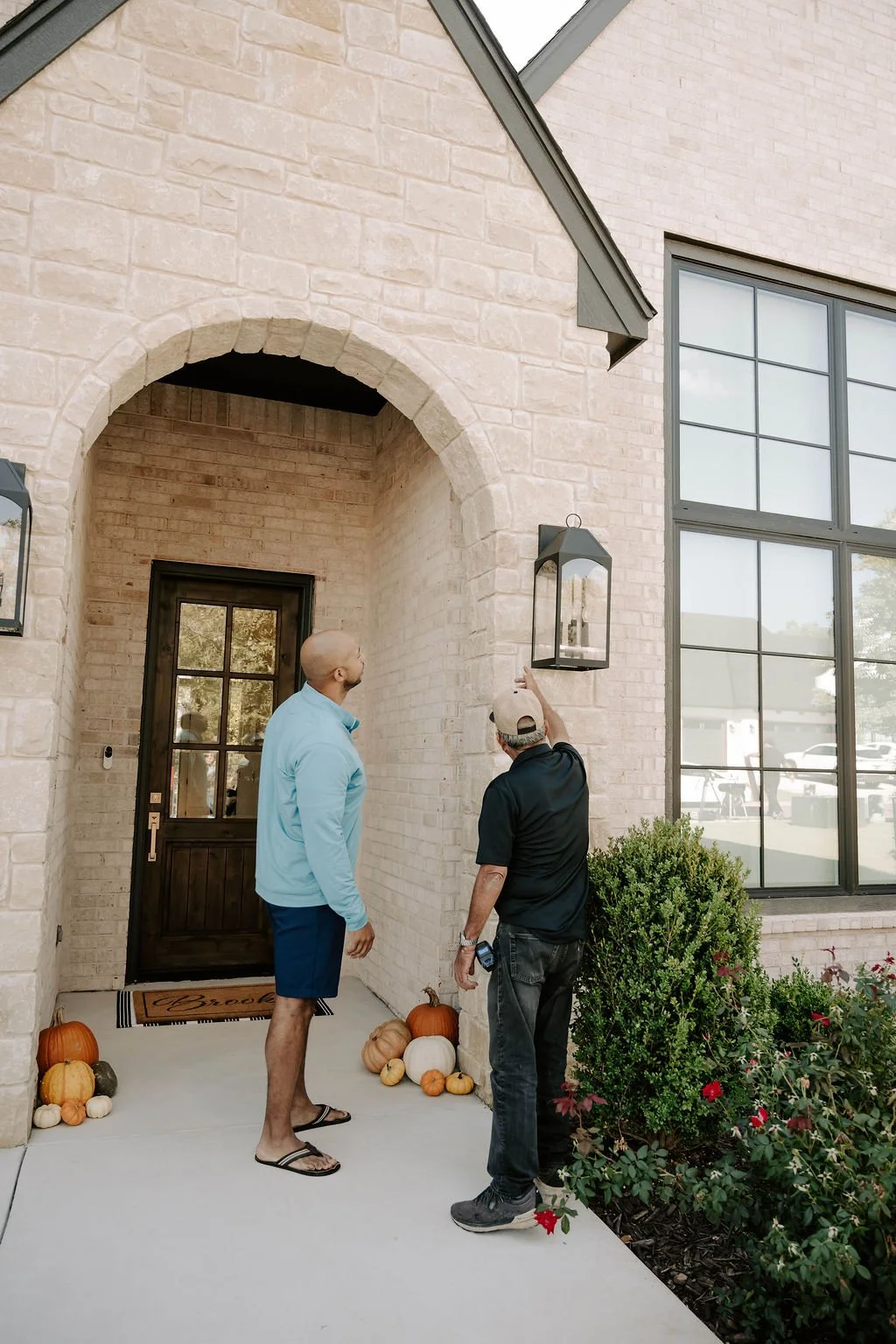Two men inspect a wall lantern outside a white brick house decorated with pumpkins.