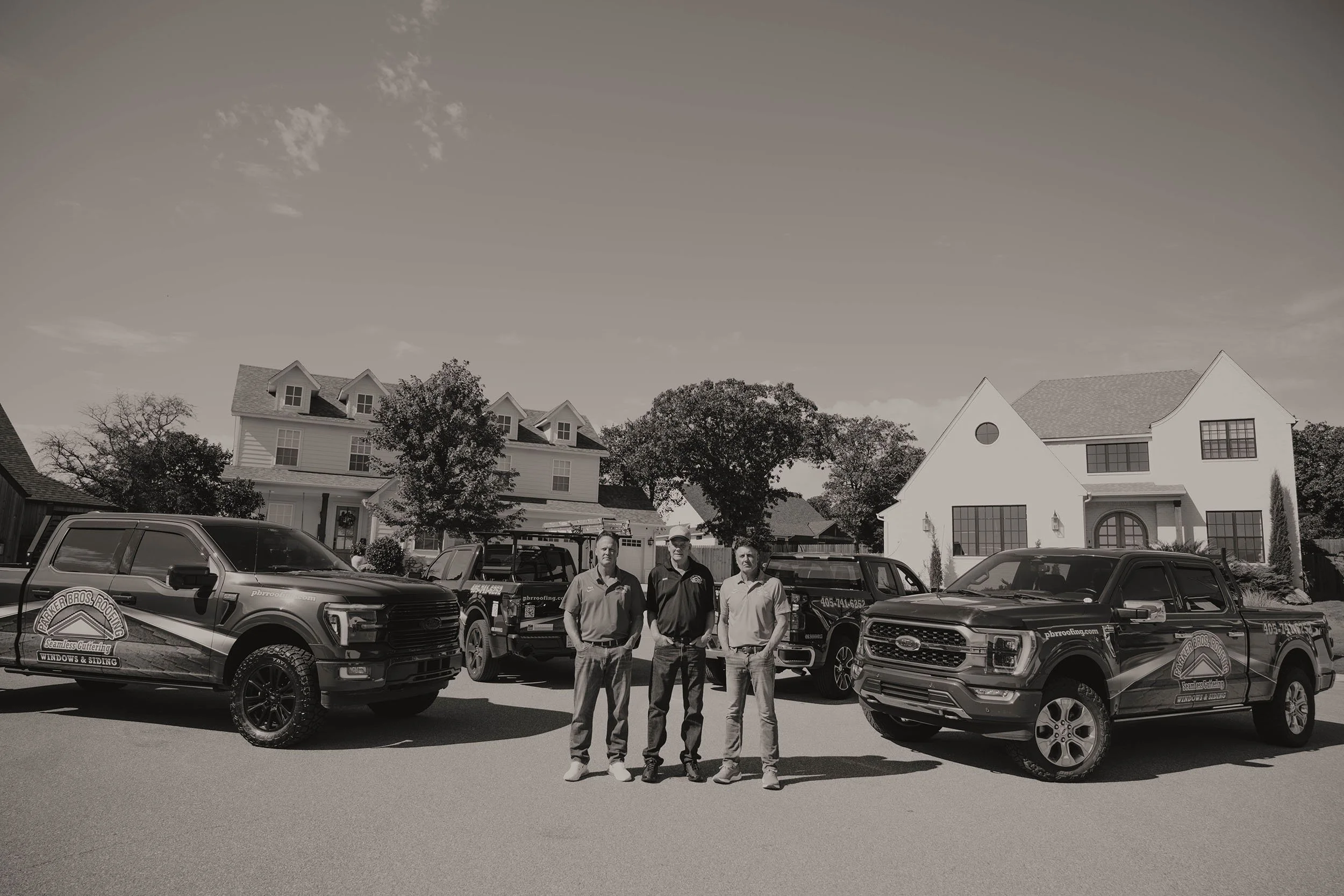 Three roofing contractors stand in front of branded work trucks and suburban homes.