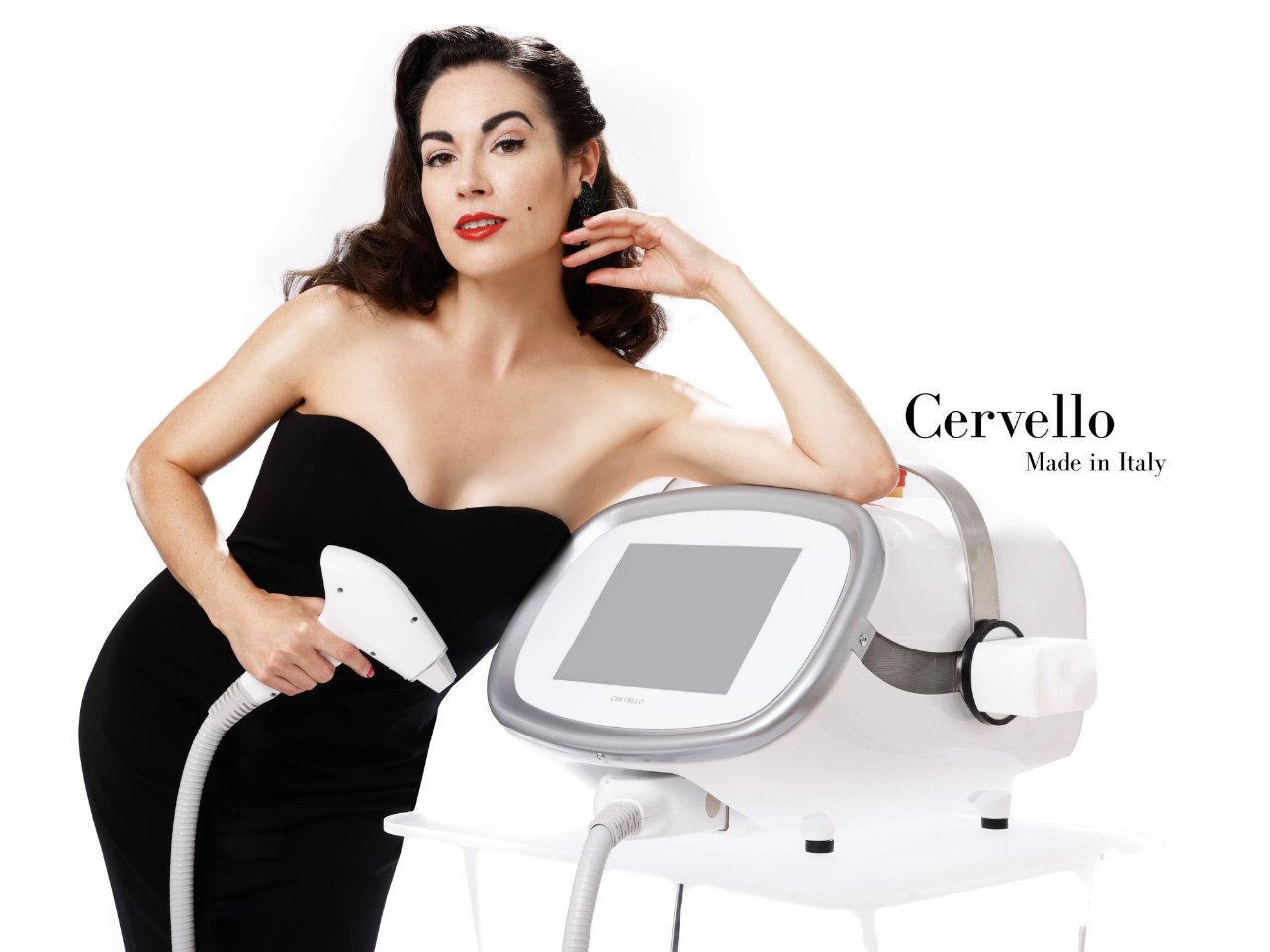 A woman with brunette hair and red lipstick wearing a black strapless dress holding a handheld beauty device, leaning on a white aesthetic machine with a large touchscreen, against a plain white background. The text "Cervello Made in Italy" is visible on the right side.