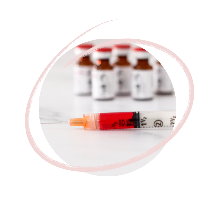 A syringe filled with red liquid in front of several vaccine vials on a white surface.