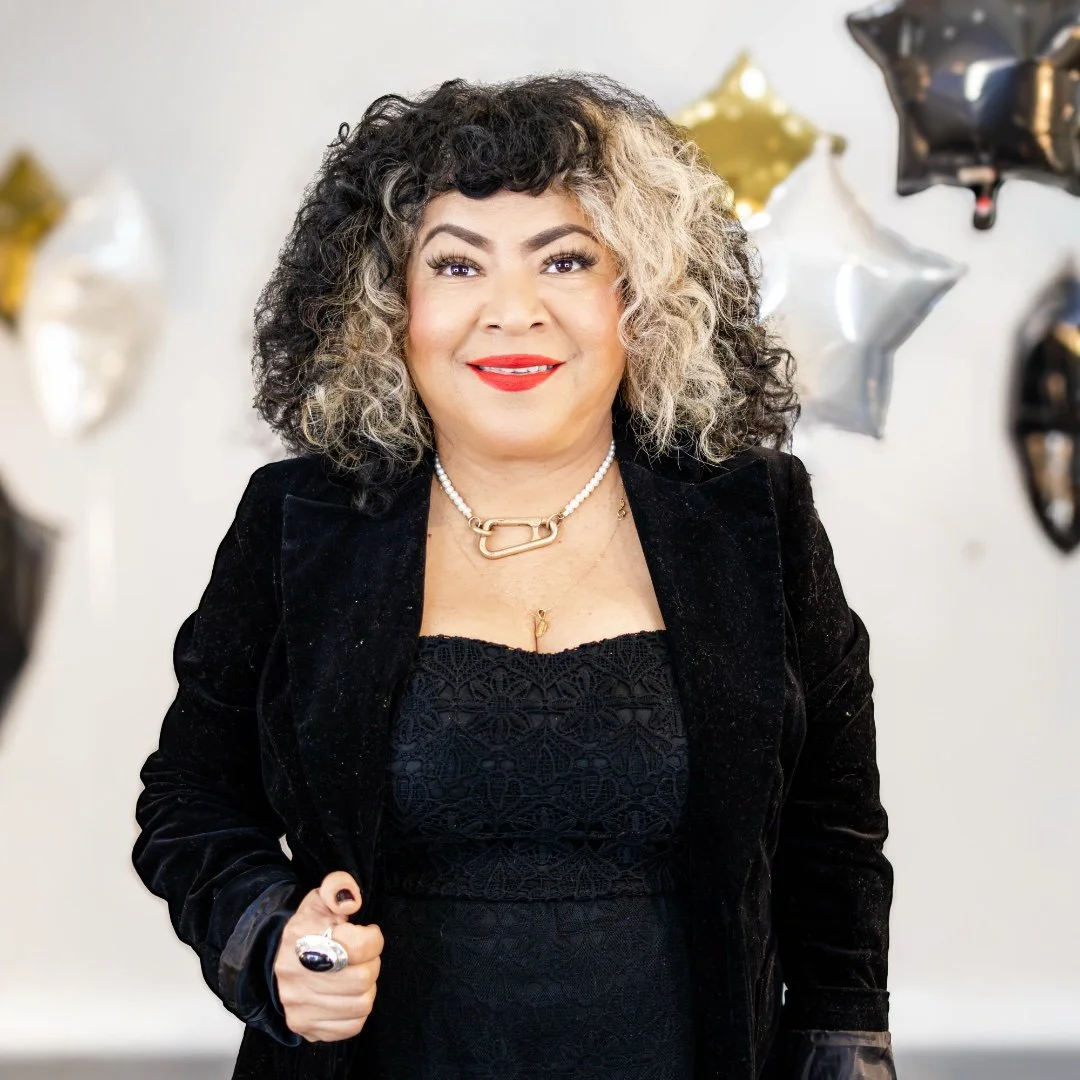 Woman with curly black and white hair, wearing a black top with rhinestones, smiling in a brightly lit salon.