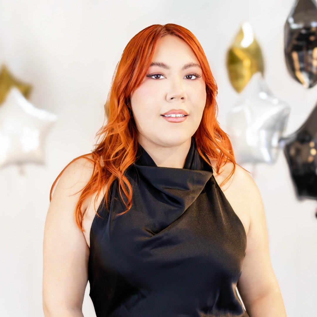 Person with red hair wearing a black top in a brightly lit room, possibly a salon.