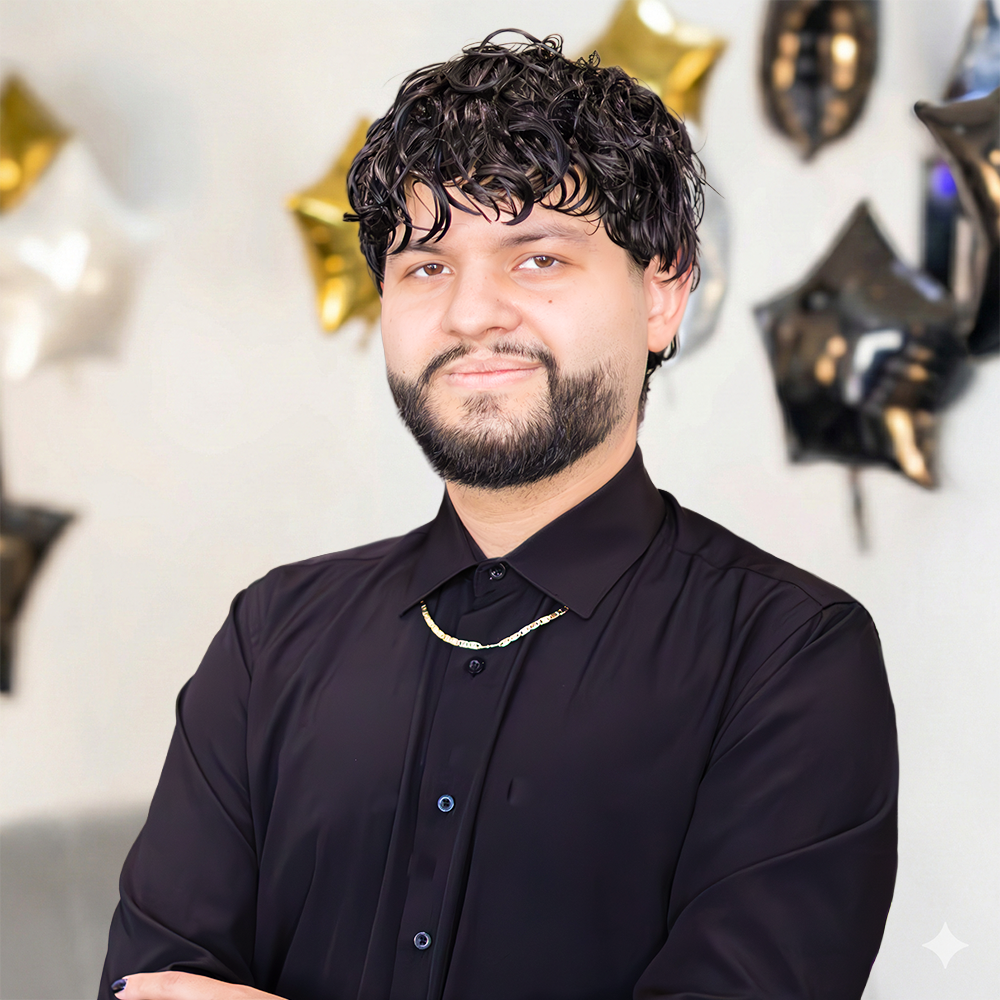 Portrait of a person with curly hair and a beard, wearing a black shirt and gold chain, smiling in an indoor setting.