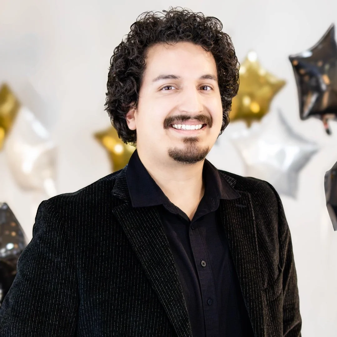 Smiling person wearing a black shirt in an indoor setting, possibly a salon or studio, with blurred background and bright lighting.