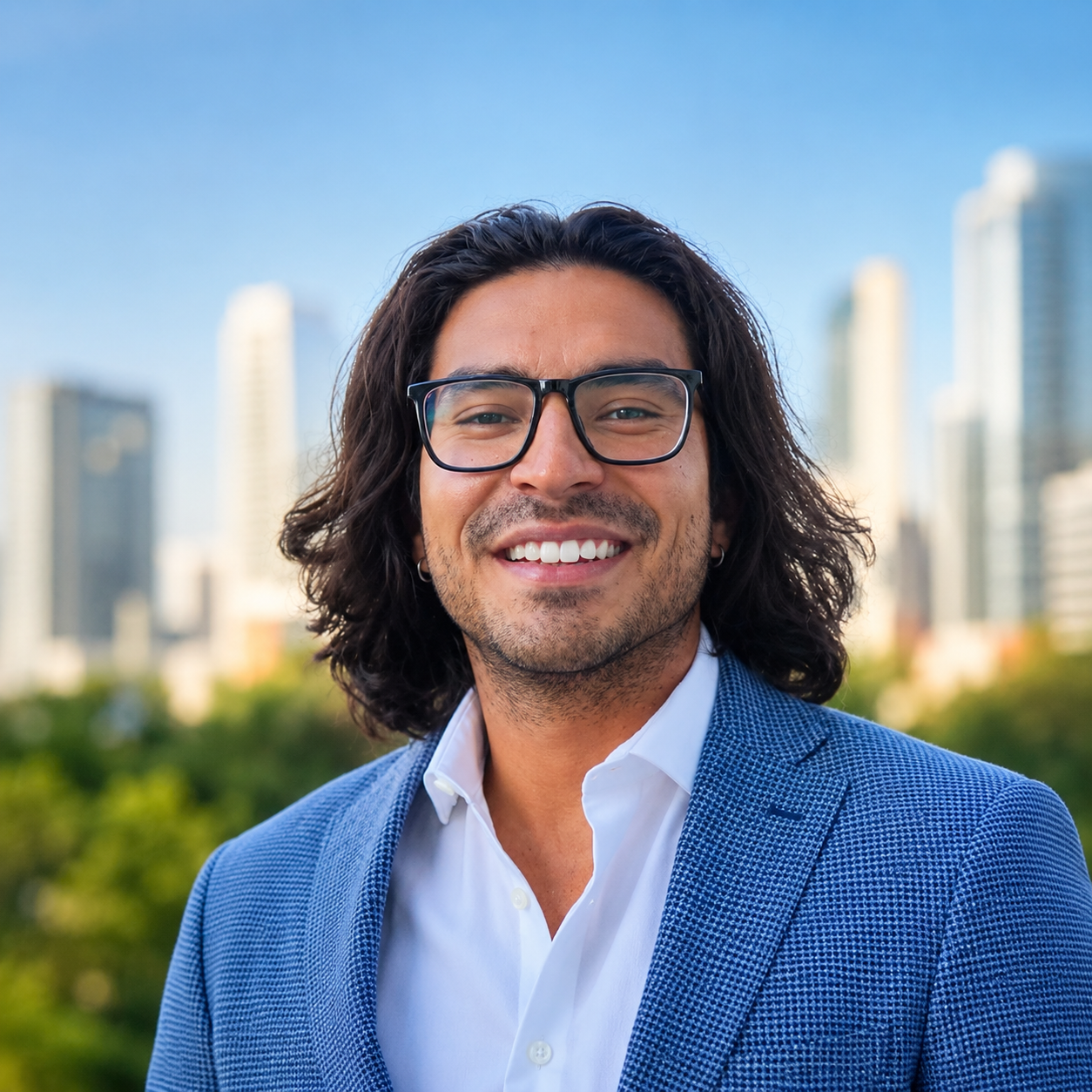 Smiling man with glasses and long, dark wavy hair dressed in a blue blazer and white shirt, with a city skyline and green trees in the background.