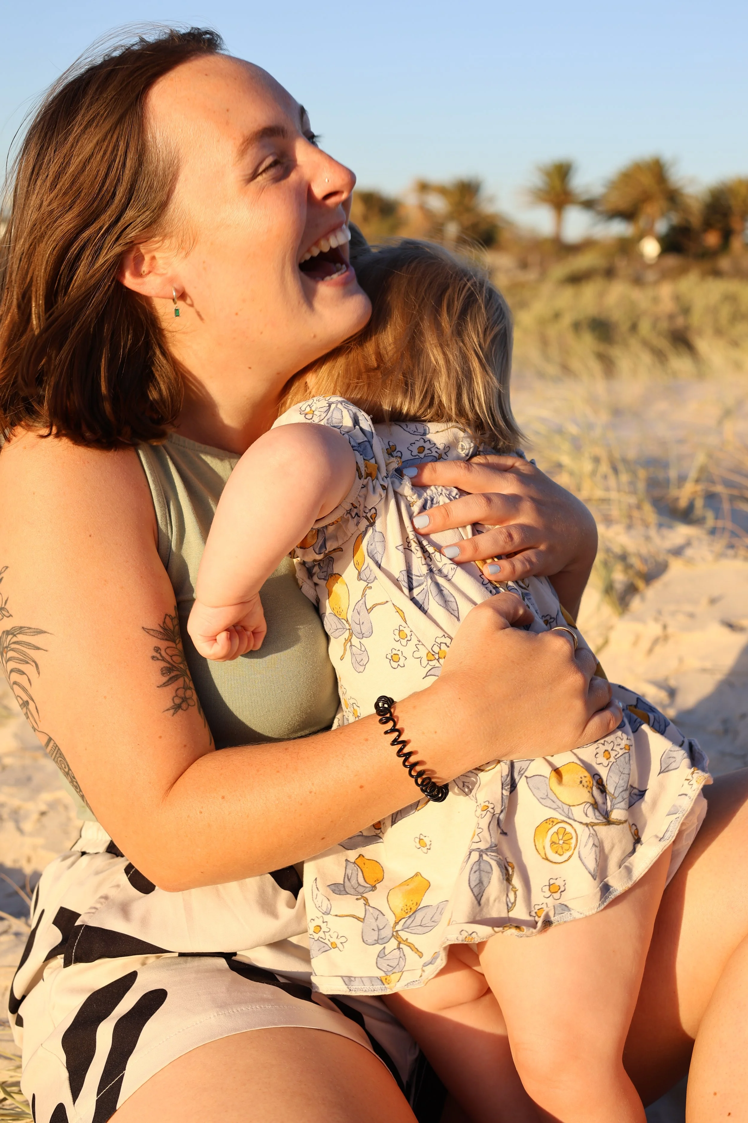 A woman and a young girl sharing a hug on a sandy beach with palm trees in the background, during sunset.