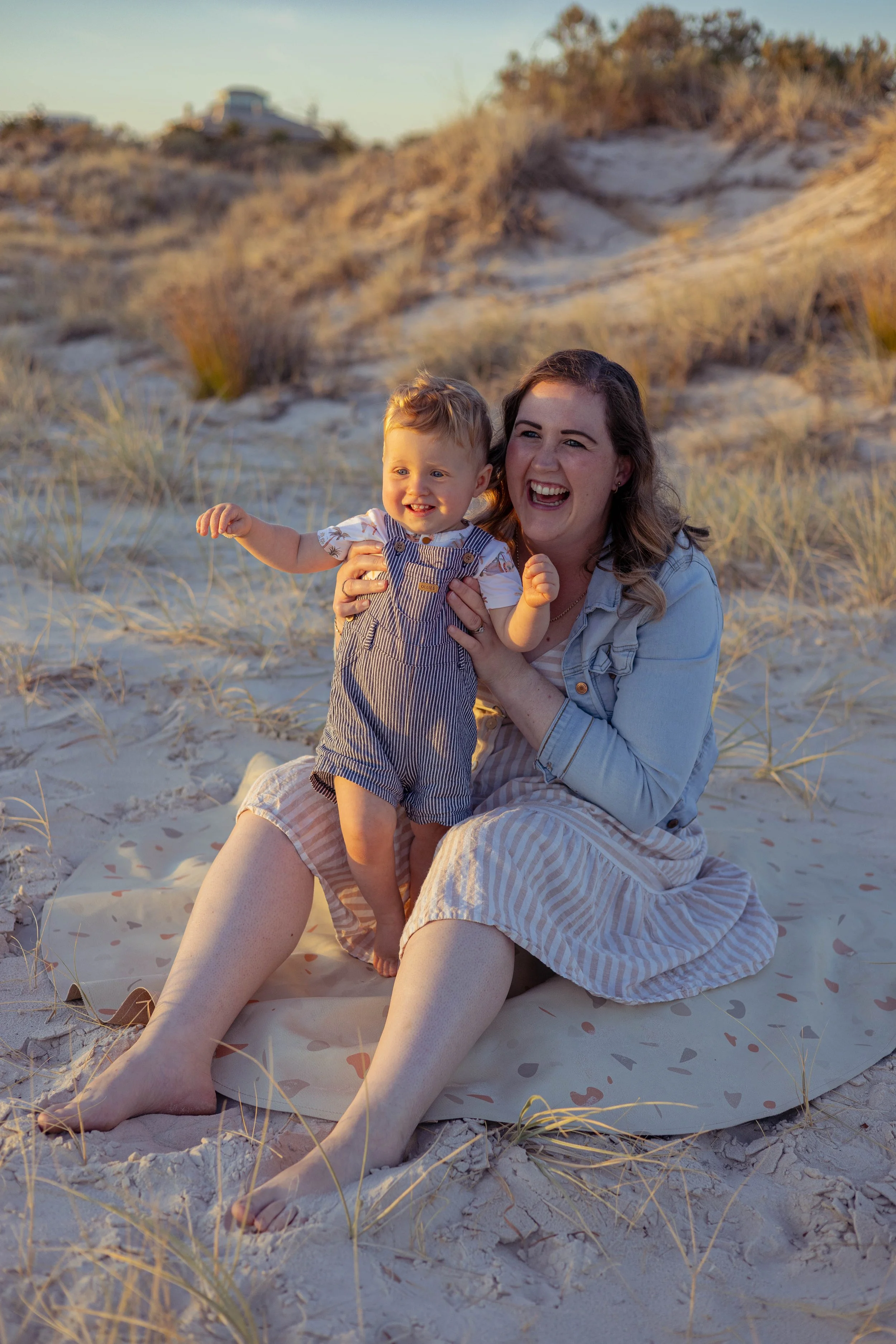 A woman and a young boy playing and smiling on a sandy beach with grassy dunes and a house in the background during sunset.