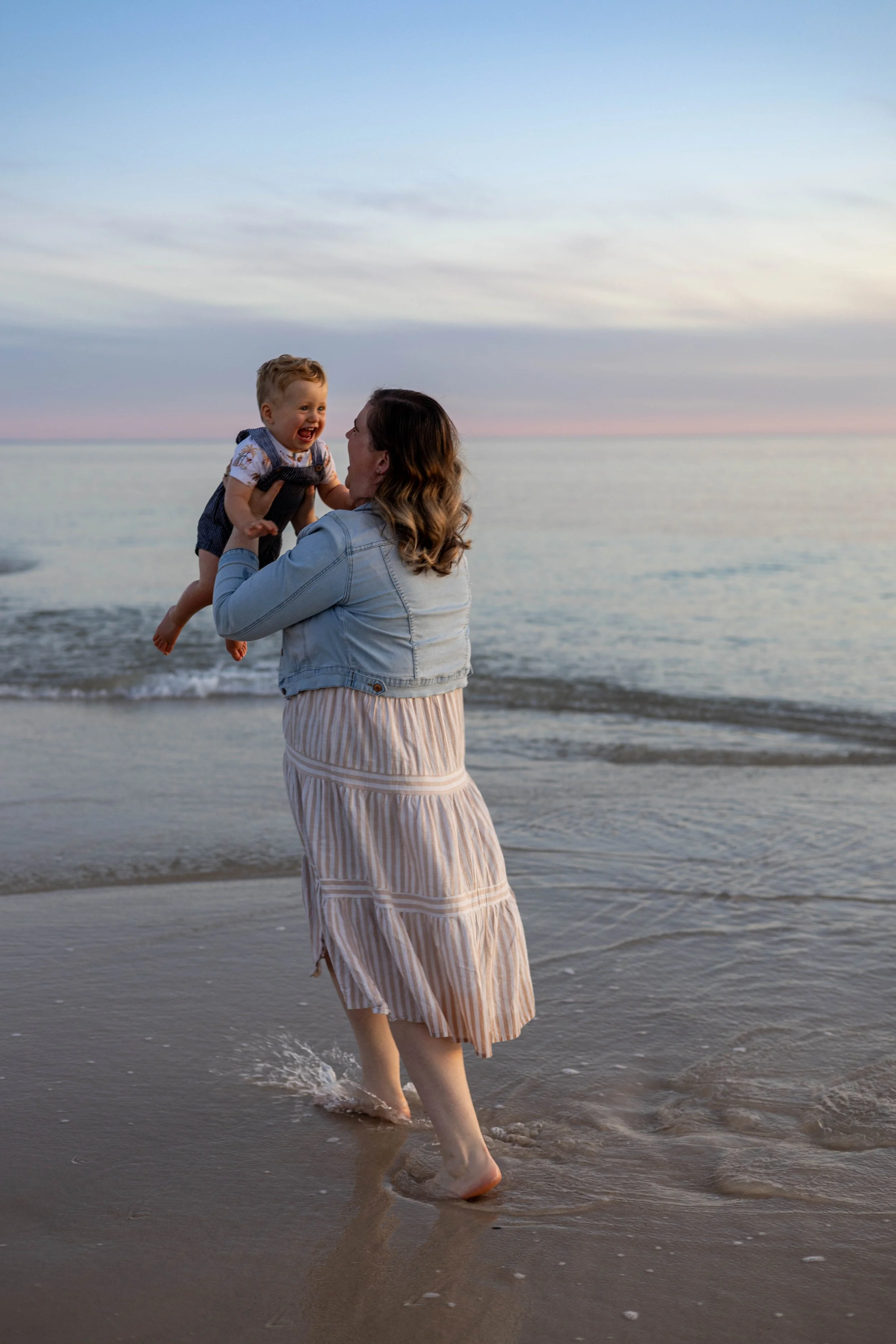 A woman playing with a young child on the beach at sunset, holding the child up in the air while the child laughs. The woman is wearing a light denim jacket and a striped dress, and the child is dressed in casual clothing.
