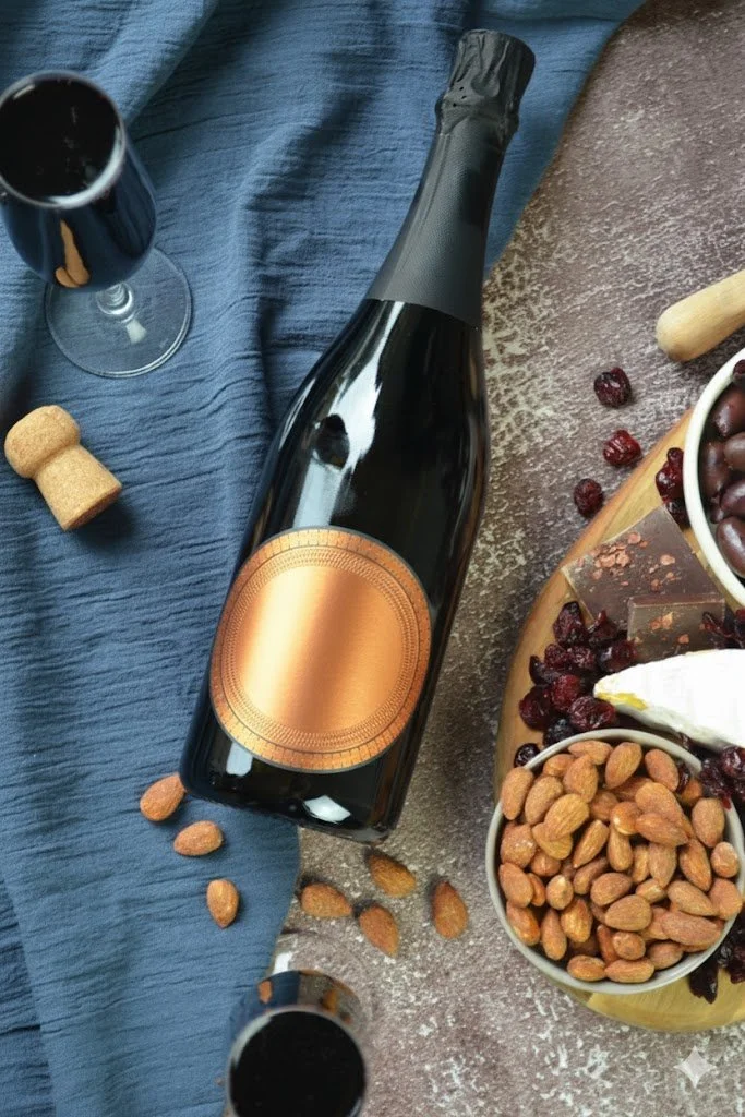 A bottle of champagne, two glasses of dark beverage, and snacks including almonds, dried cranberries, and chocolates on a table with a blue cloth and a brown textured surface.