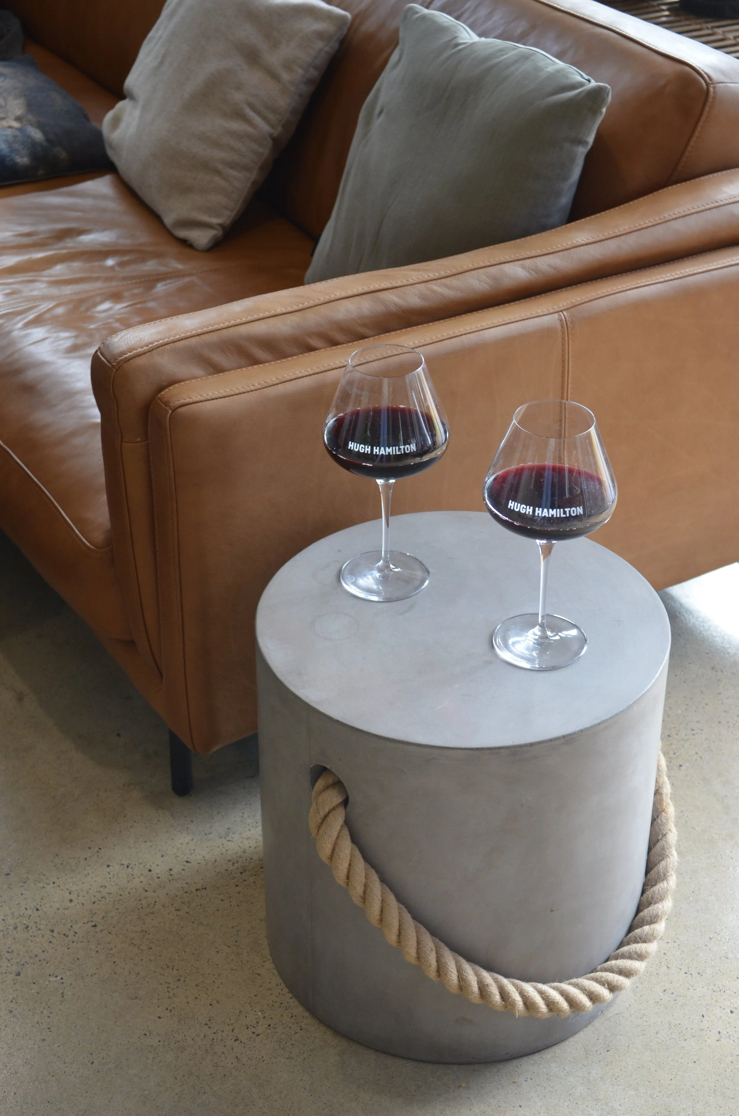 Two glasses of red wine on a round concrete side table with a rope handle, in front of a tan leather sofa with beige and green cushions.