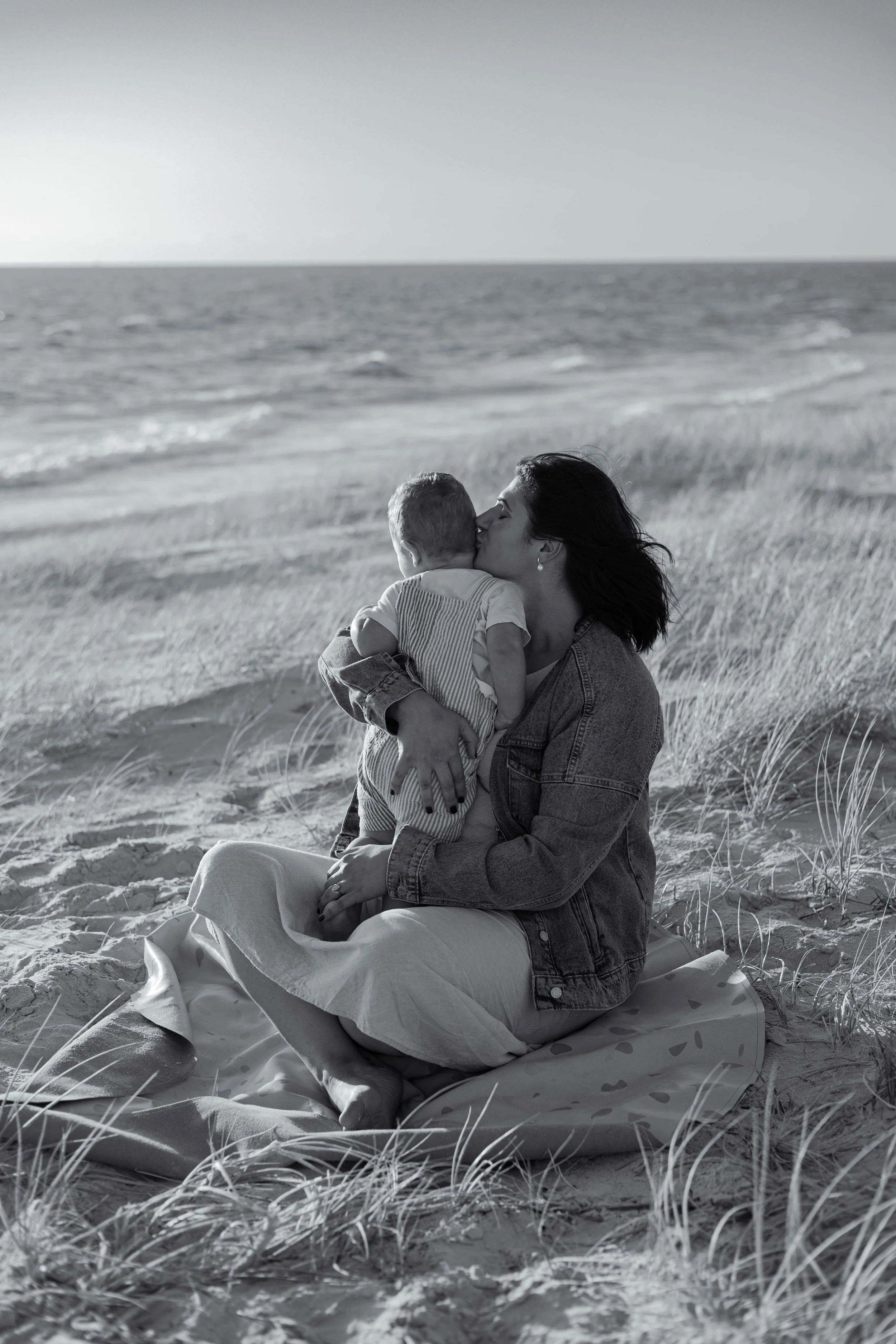 A woman and a young child sharing a kiss on a blanket on the beach with dunes and ocean in the background.
