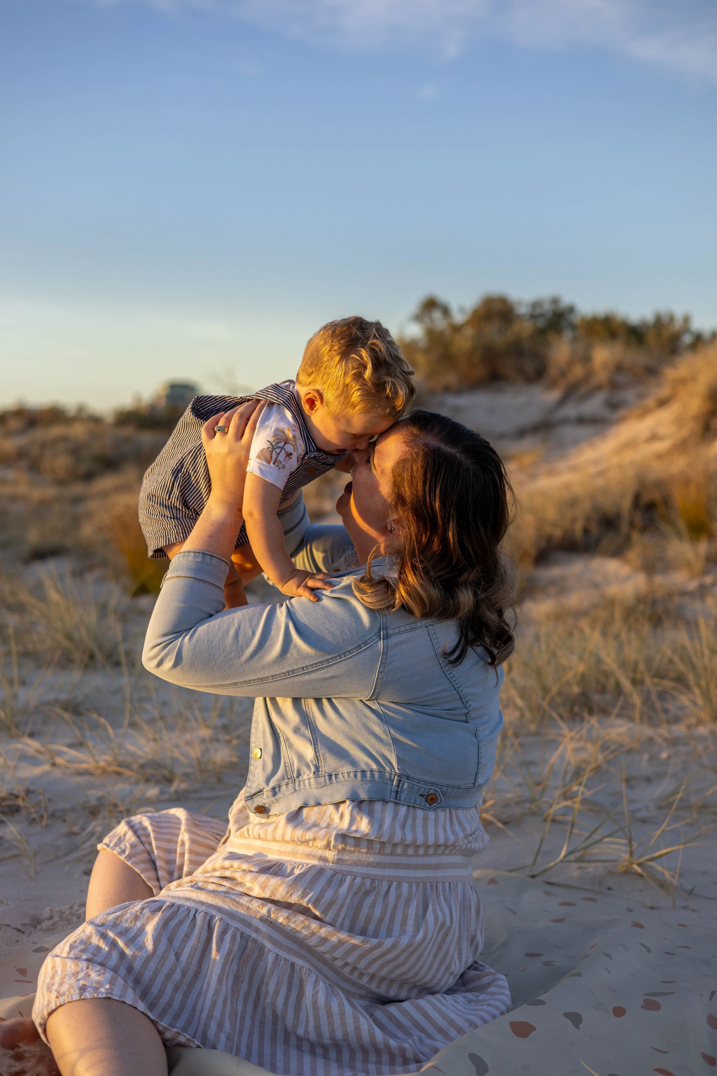 A woman sitting on a blanket at the beach holding a baby up in the air, touching foreheads in a kiss during sunset.