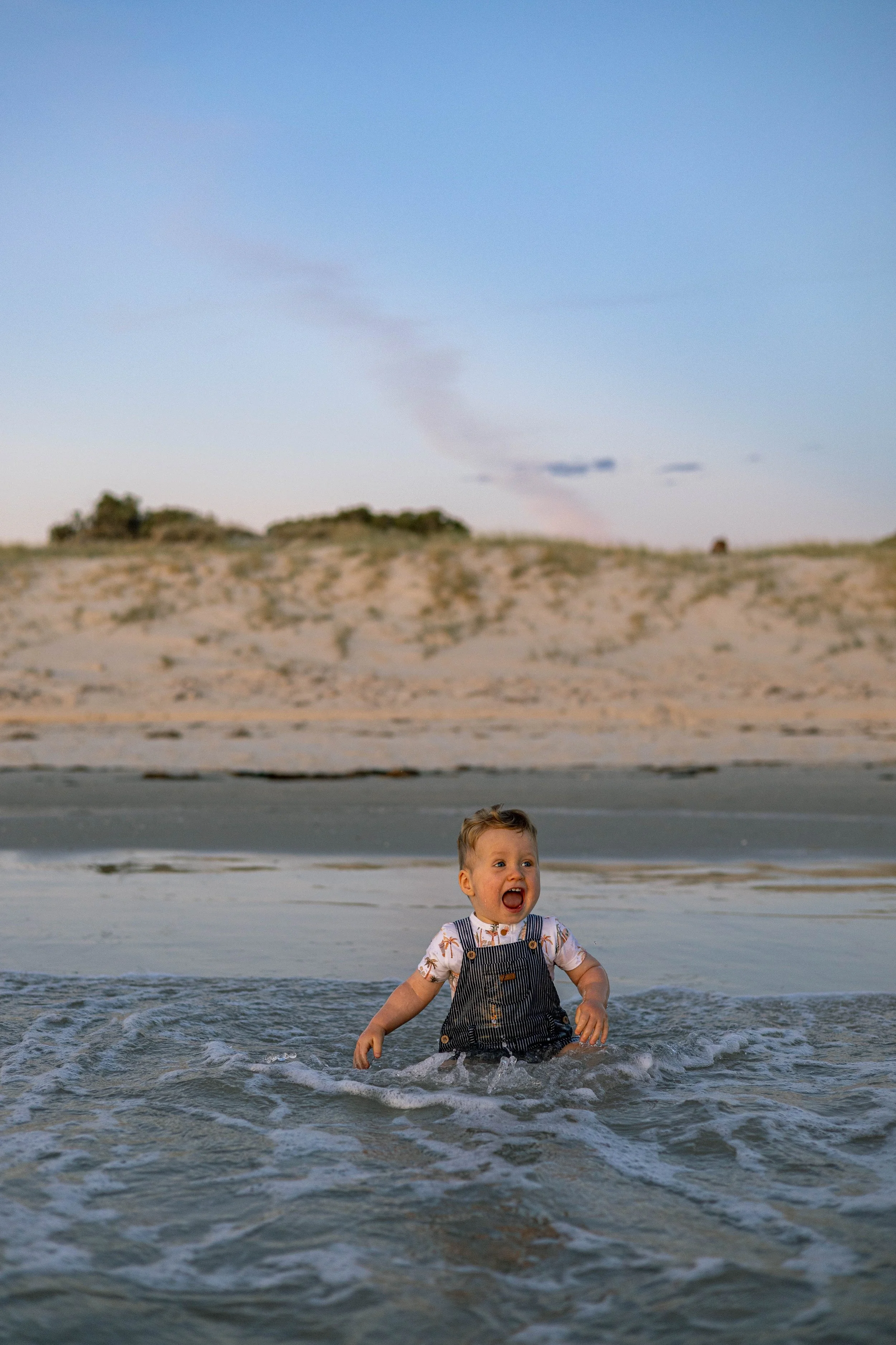 A young boy in striped overalls playing in the ocean water at the beach with a sandy dune and sky in the background.