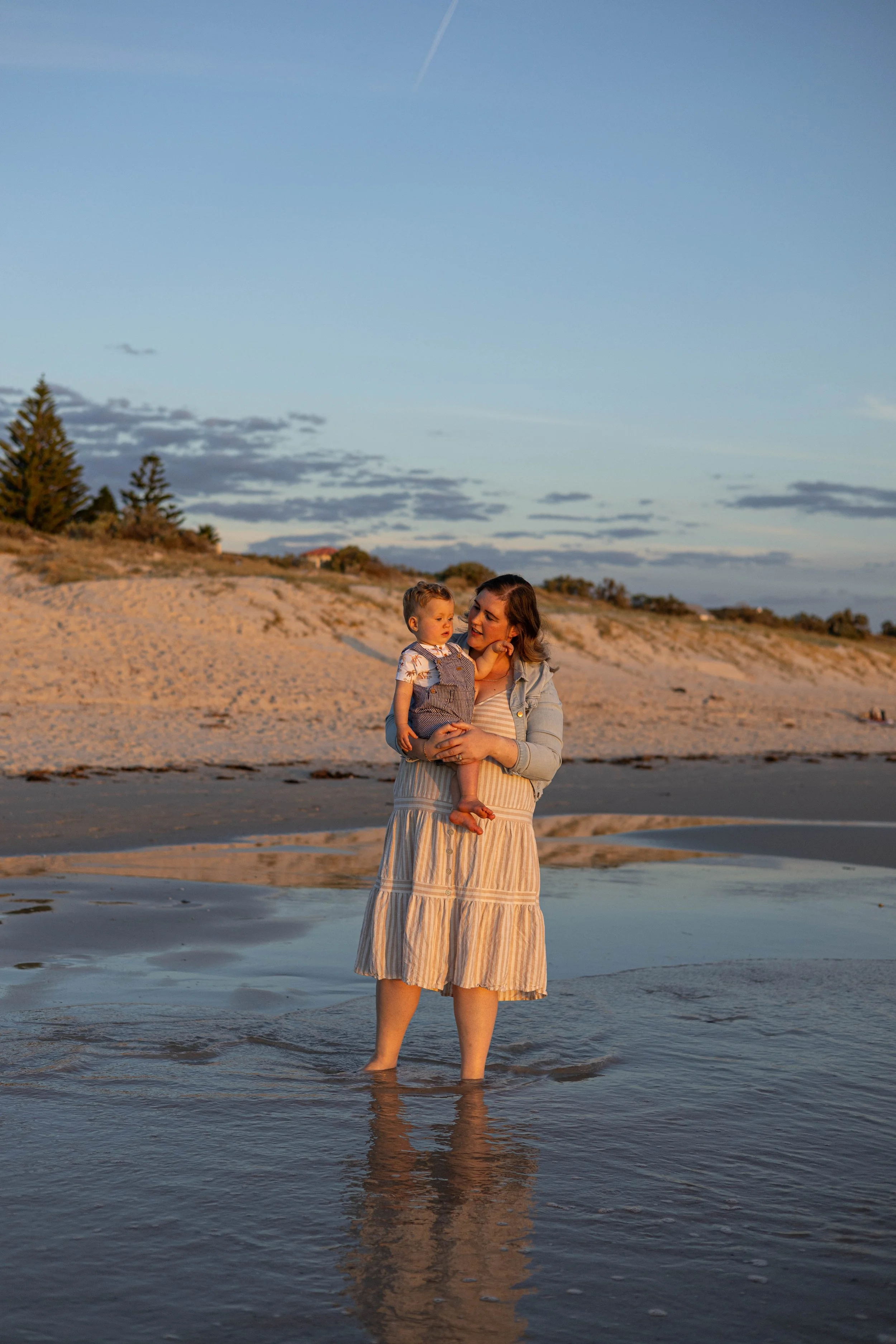 A woman and a child stand in shallow water on a beach during sunset, with sand dunes and trees in the background.