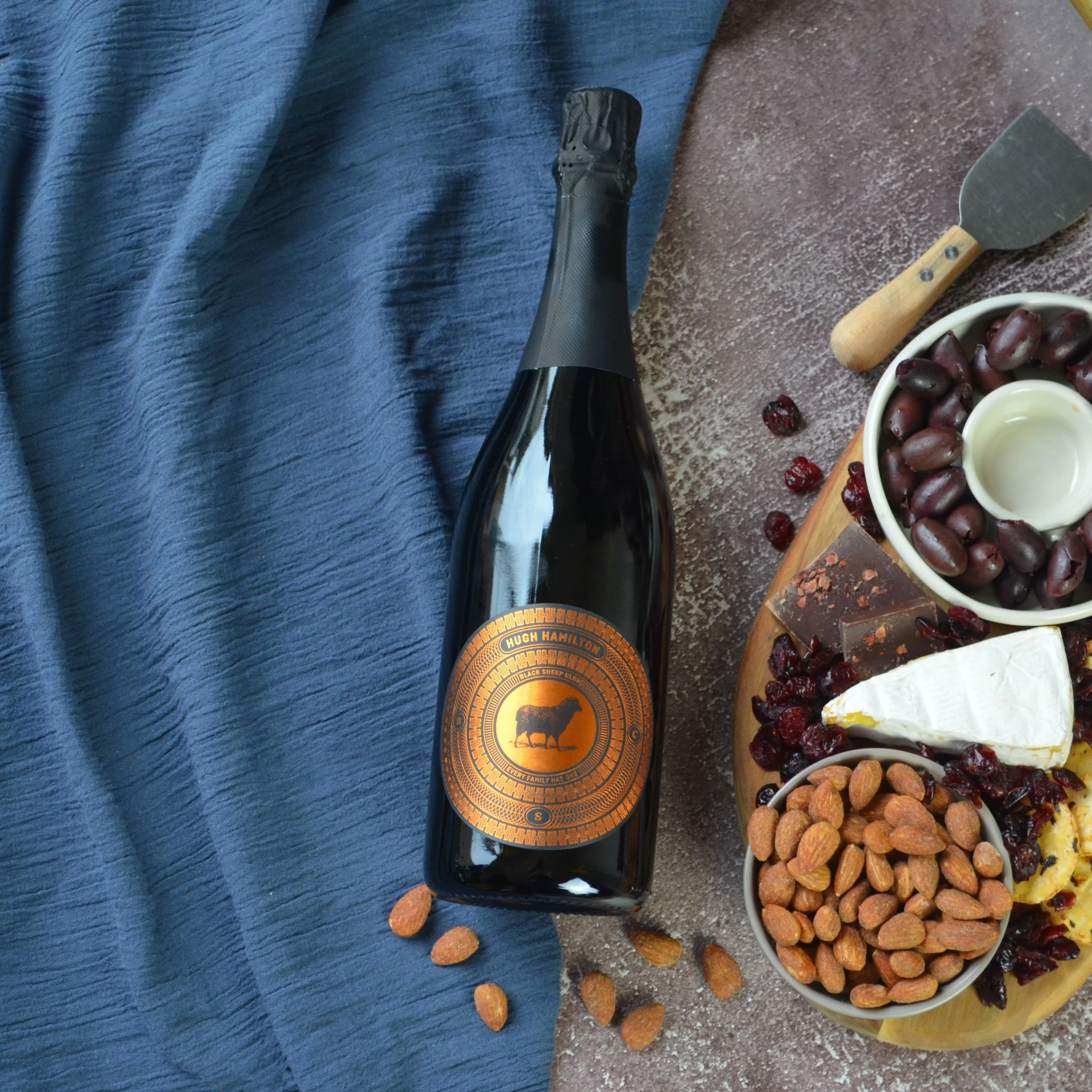 A bottle of sparkling wine or champagne with a dark label, lying on a blue fabric. Next to it is a cheese and fruit platter containing grapes, dried apricots, and dried cranberries, with a small cheese wedge and some chocolates. There is also a chees