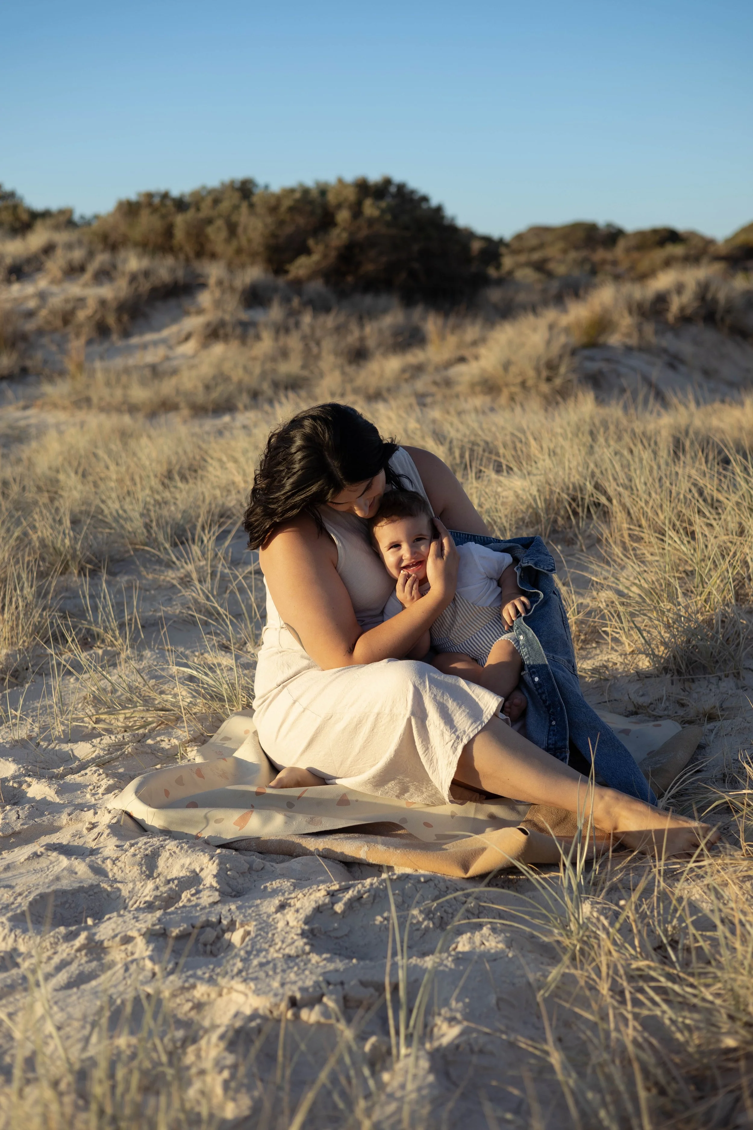 A woman and a young boy sitting on the sandy beach with tall grass, embracing and smiling during sunset or late afternoon.
