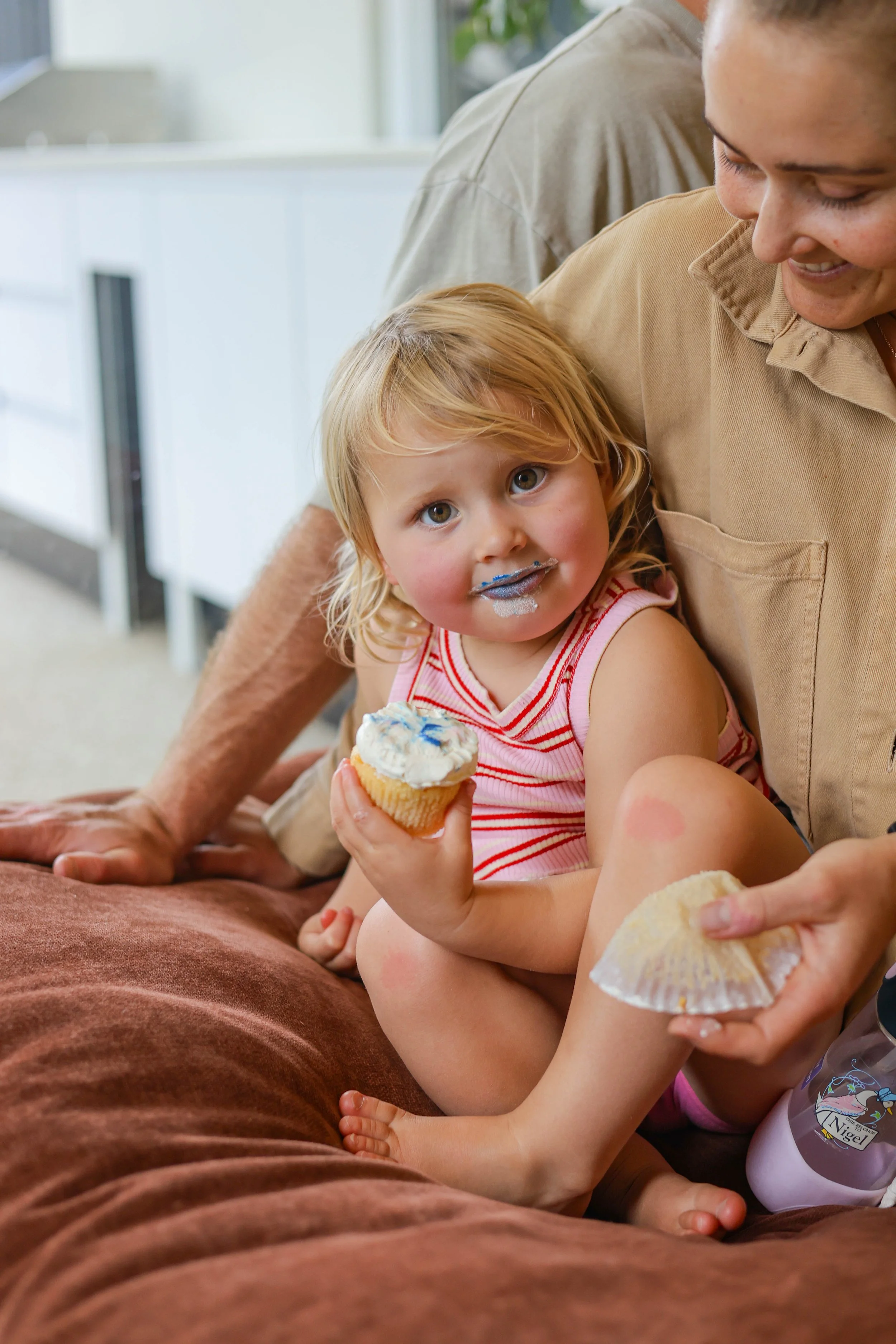 Young girl with blonde hair, wearing a pink and white striped tank top, sitting on a brown couch holding a cupcake with white frosting and blue sprinkles. She has blue icing on her face around her mouth. An adult woman, smiling, is near her, holding 