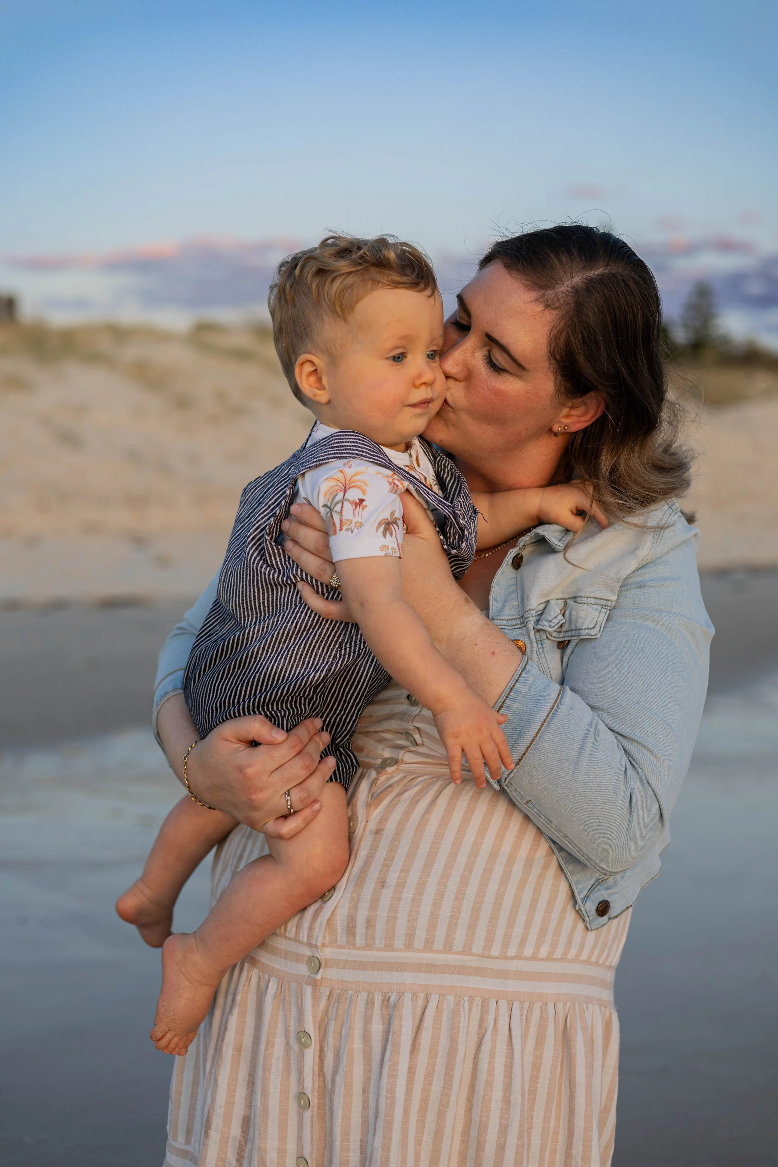 A woman holding a young boy at the beach during sunset, sharing an affectionate moment.