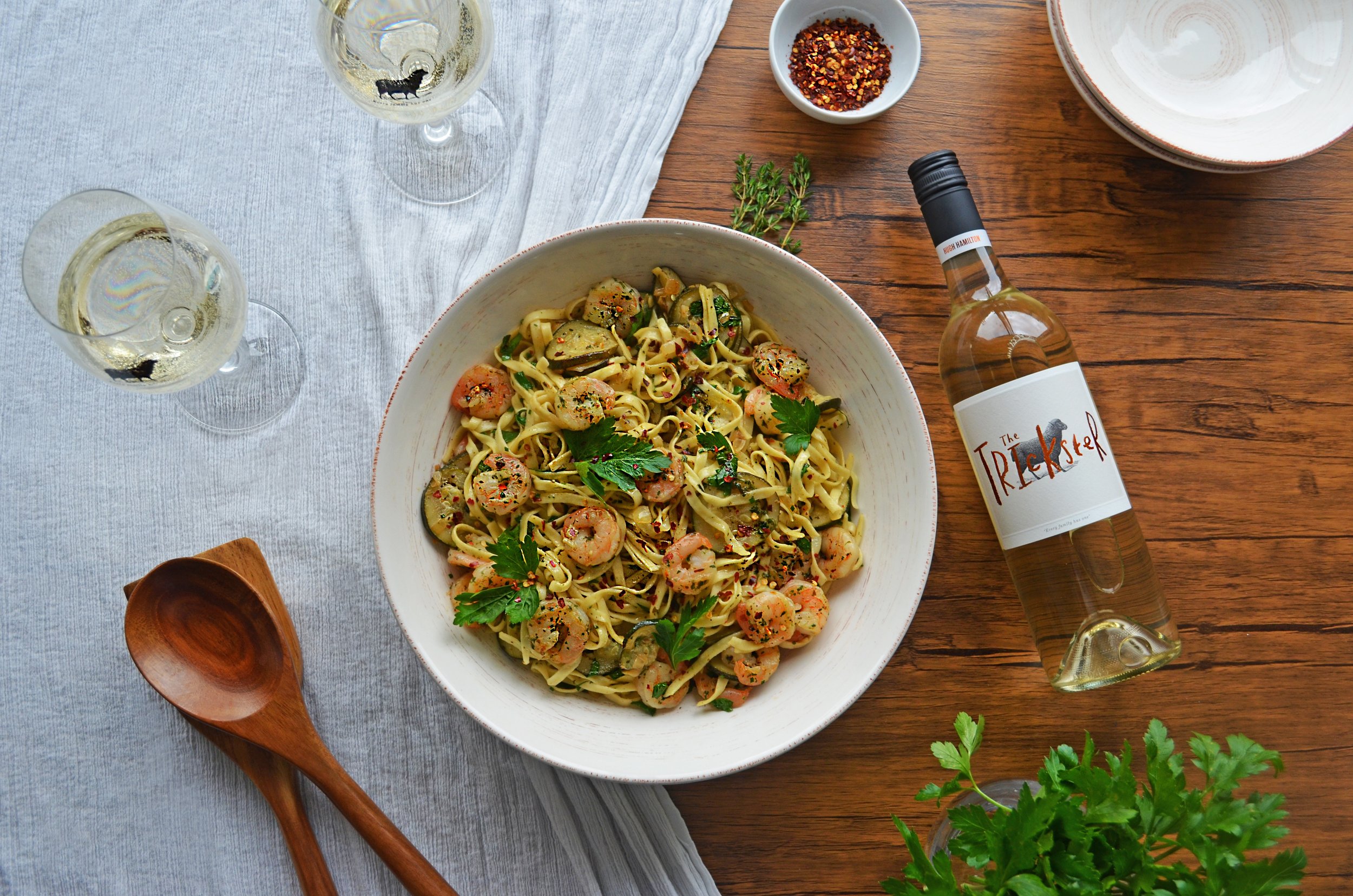 A bowl of prawns and pasta garnished with fresh parsley, served on a wooden table with white wine, a bottle, a small bowl of crushed red pepper, and fresh herbs.