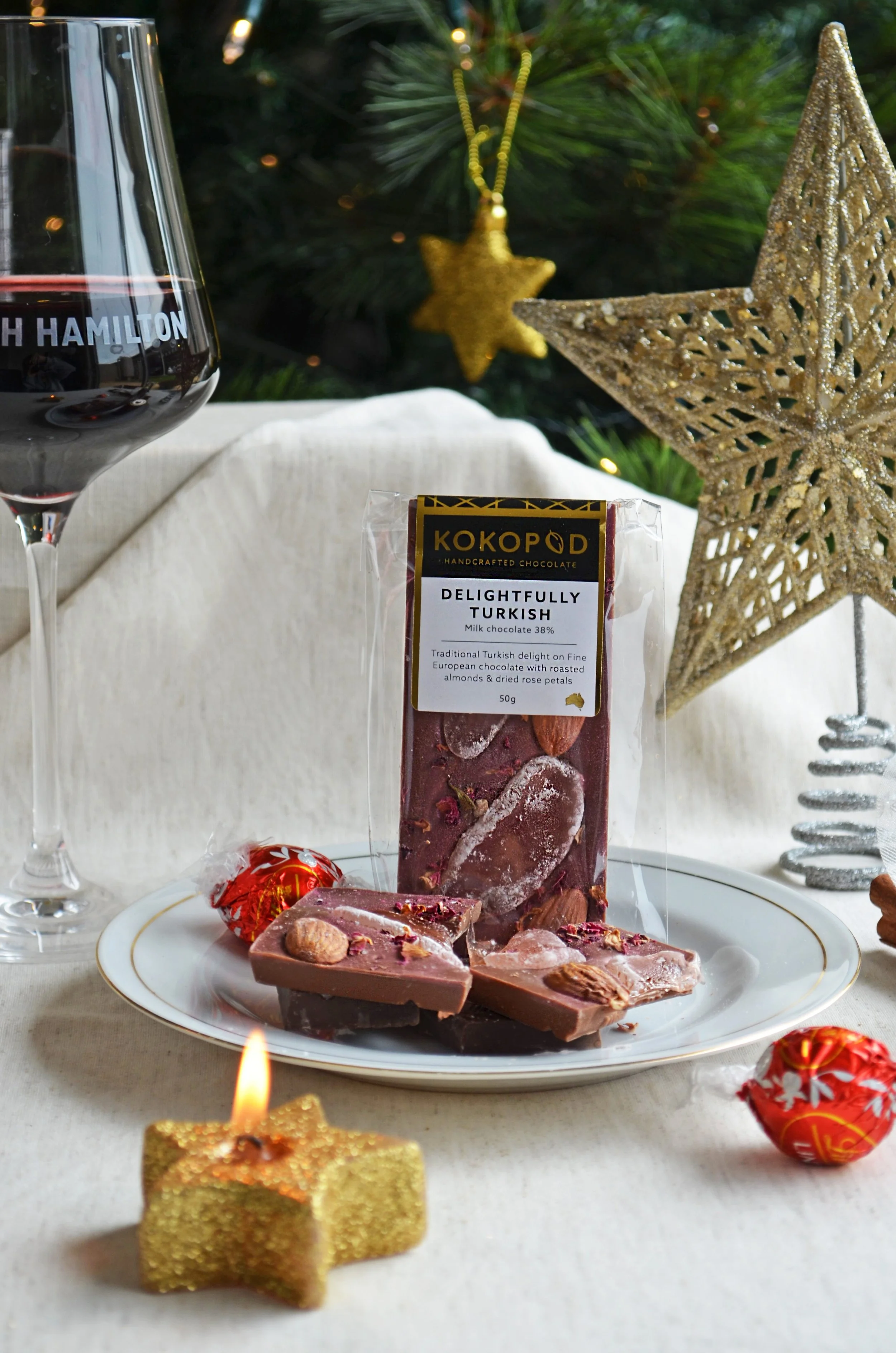 A plate of handcrafted Turkish milk chocolate with almonds and rose petals, surrounded by Christmas decorations including a gold star candle, a large gold star ornament, a glass of red wine, and wrapped candies, with a Christmas tree in the backgroun
