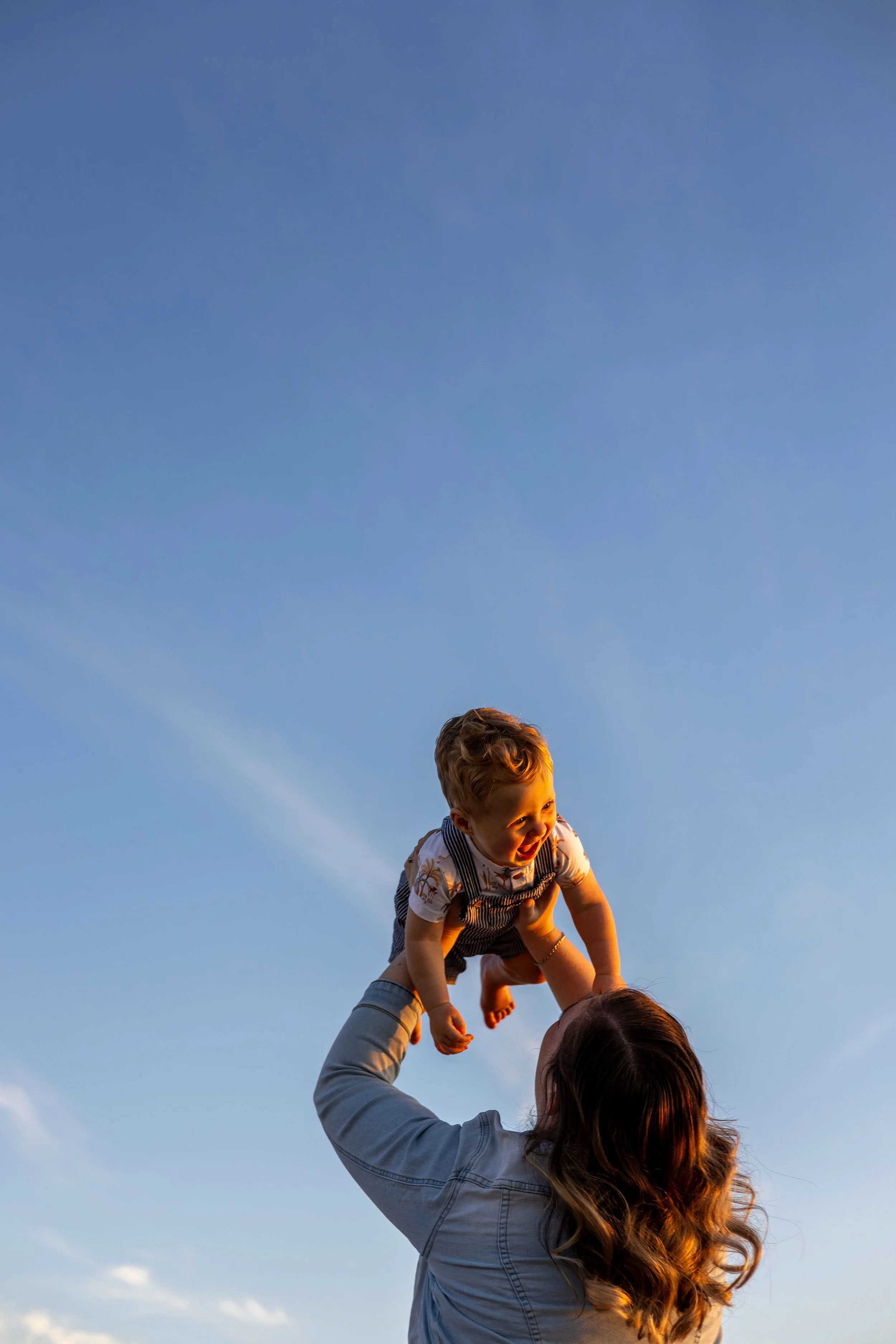 A woman lifting a young boy into the air outside during sunset, with a clear blue sky in the background.