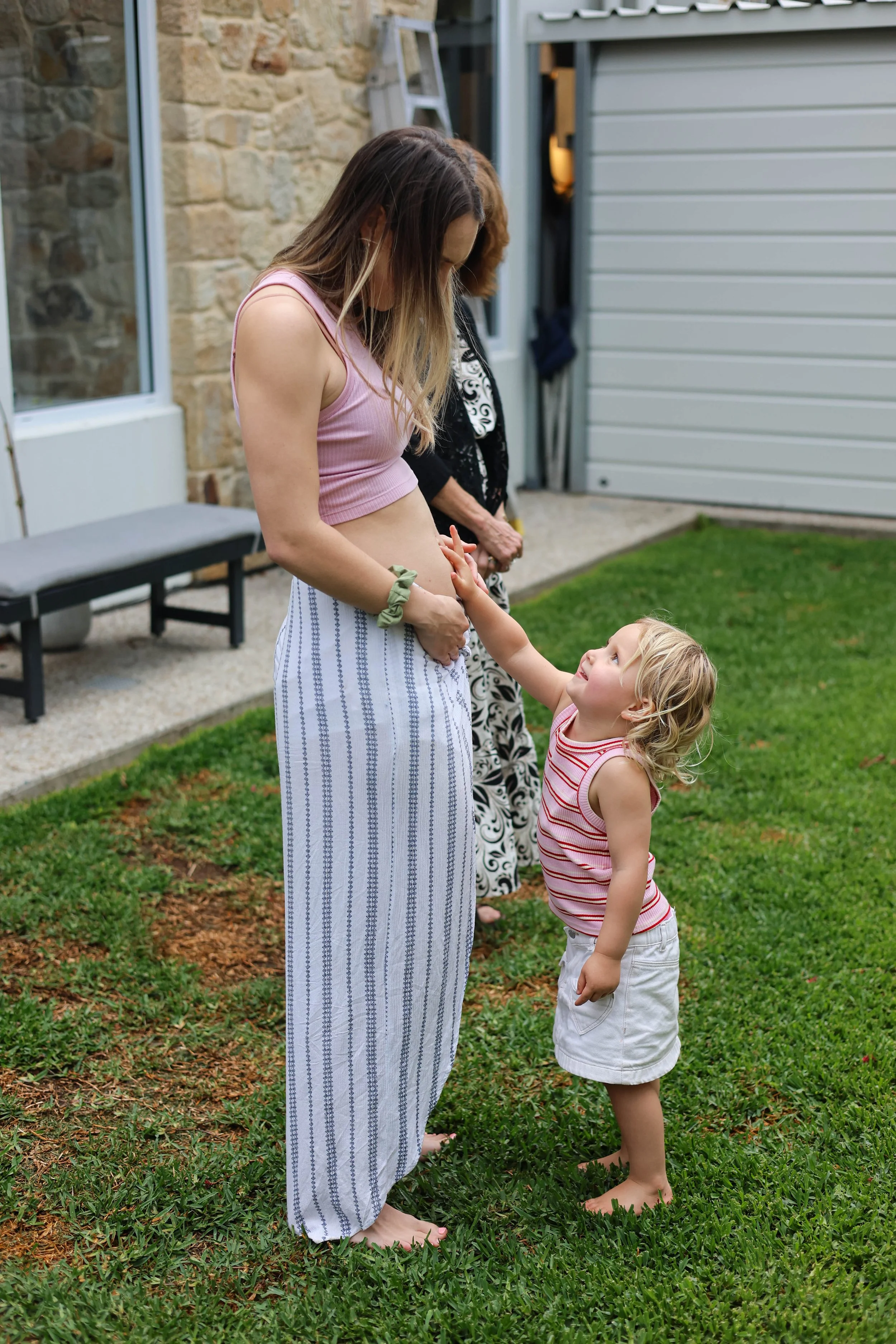 A young girl touching a pregnant woman's belly, smiling, outdoors on a grassy area with a stone wall and a garage in the background, and an older woman standing behind them.