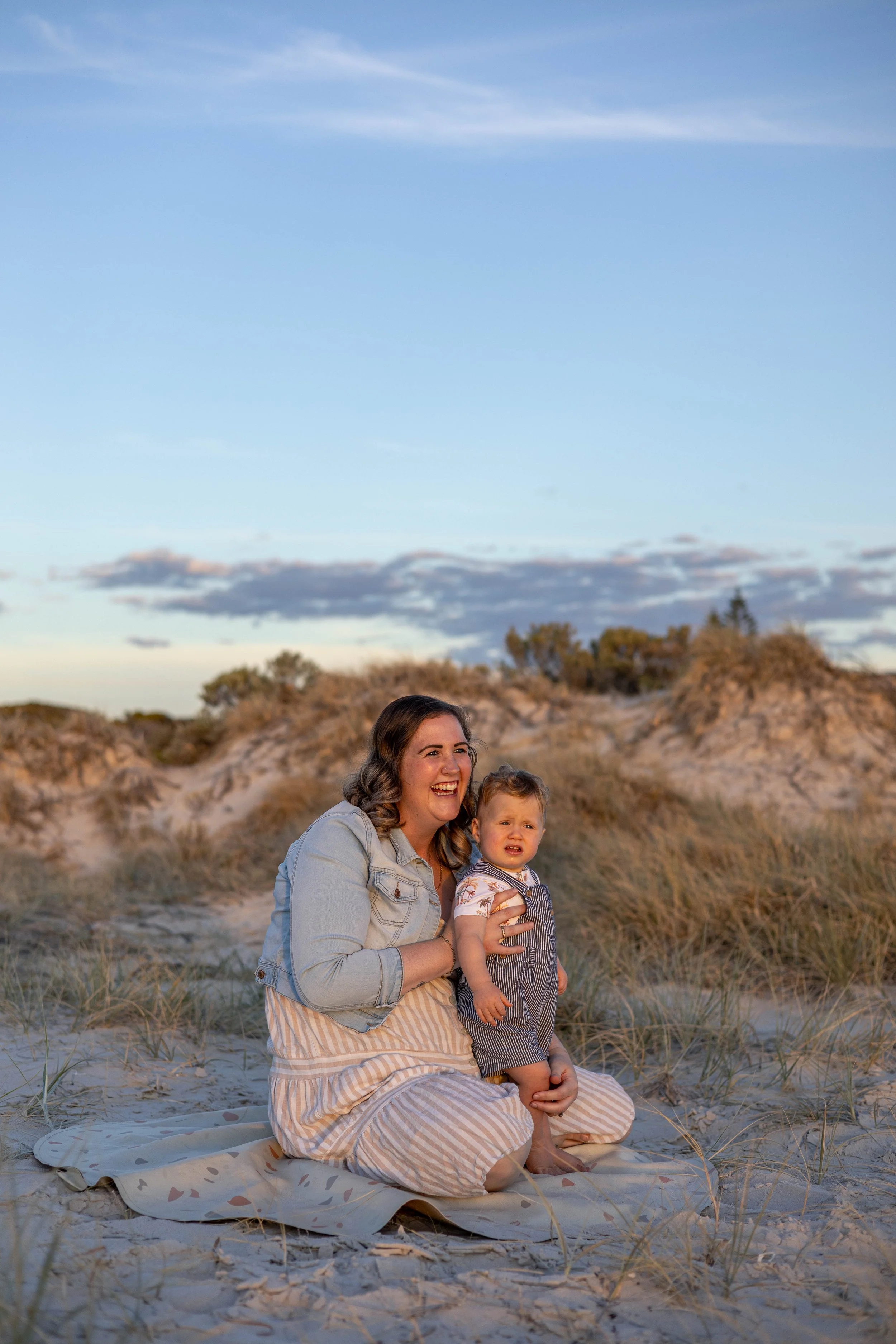 A woman and a young boy sitting on a blanket at the beach during sunset, with sand dunes and sky in the background.