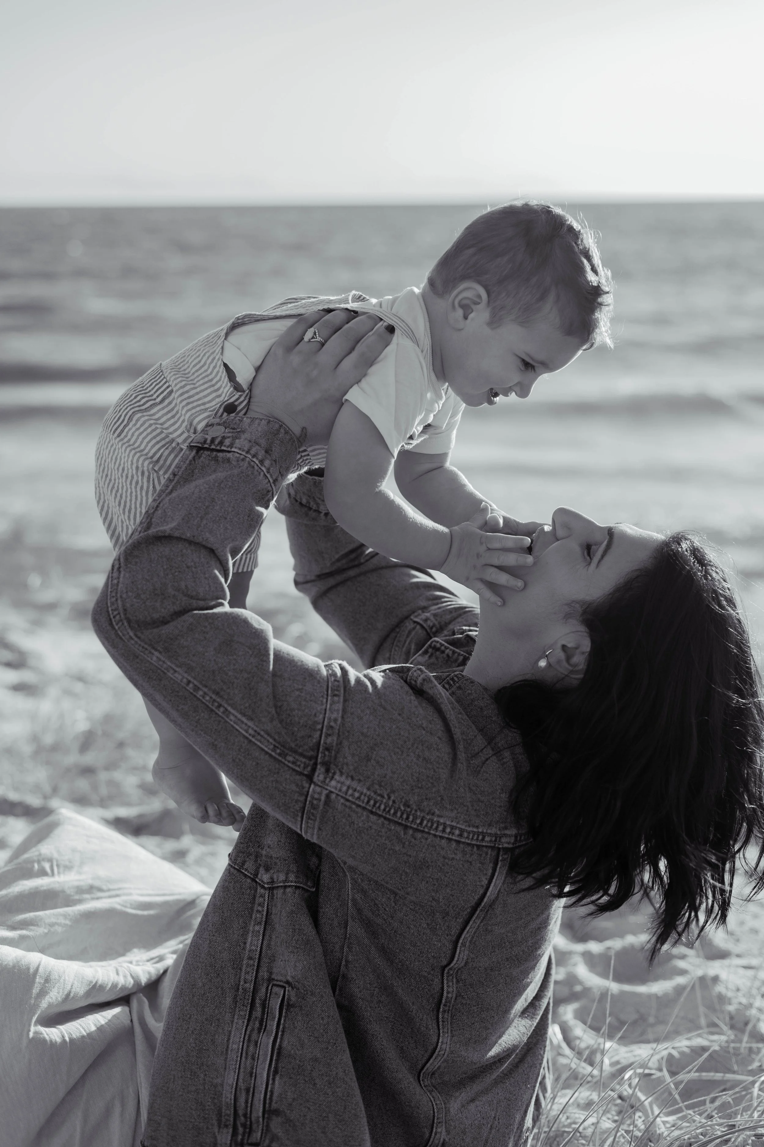 A woman playing with a young child on a beach, holding the child up in the air, both smiling and enjoying their time together.