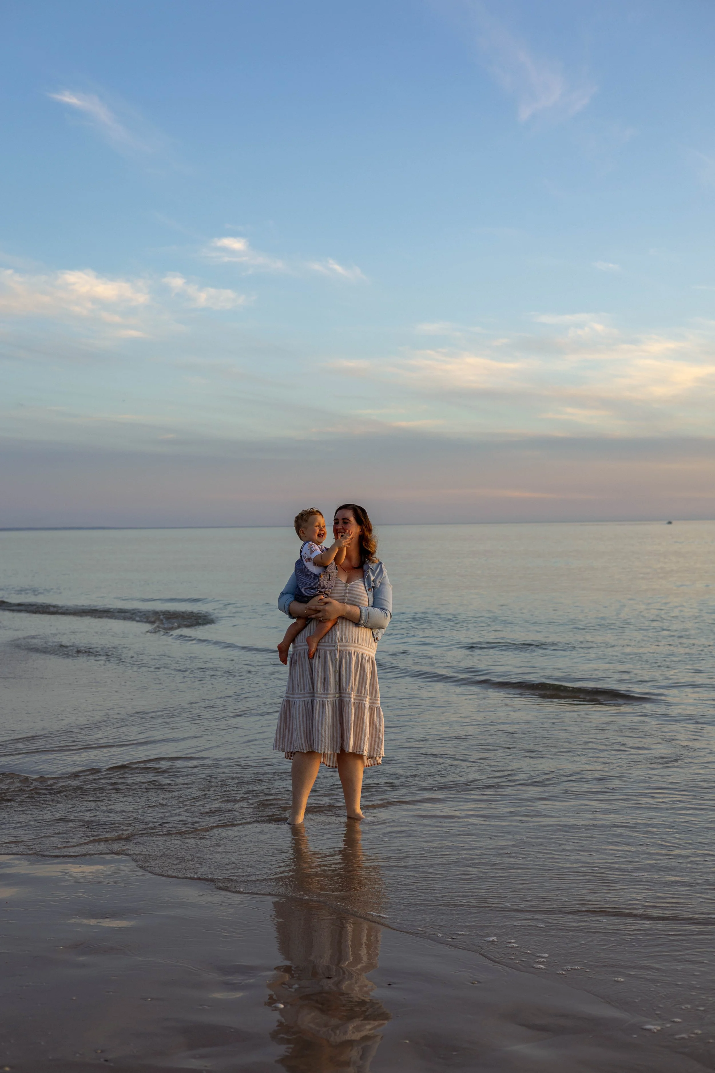 A woman holding a young child on a beach during sunset, with the ocean and sky in the background.