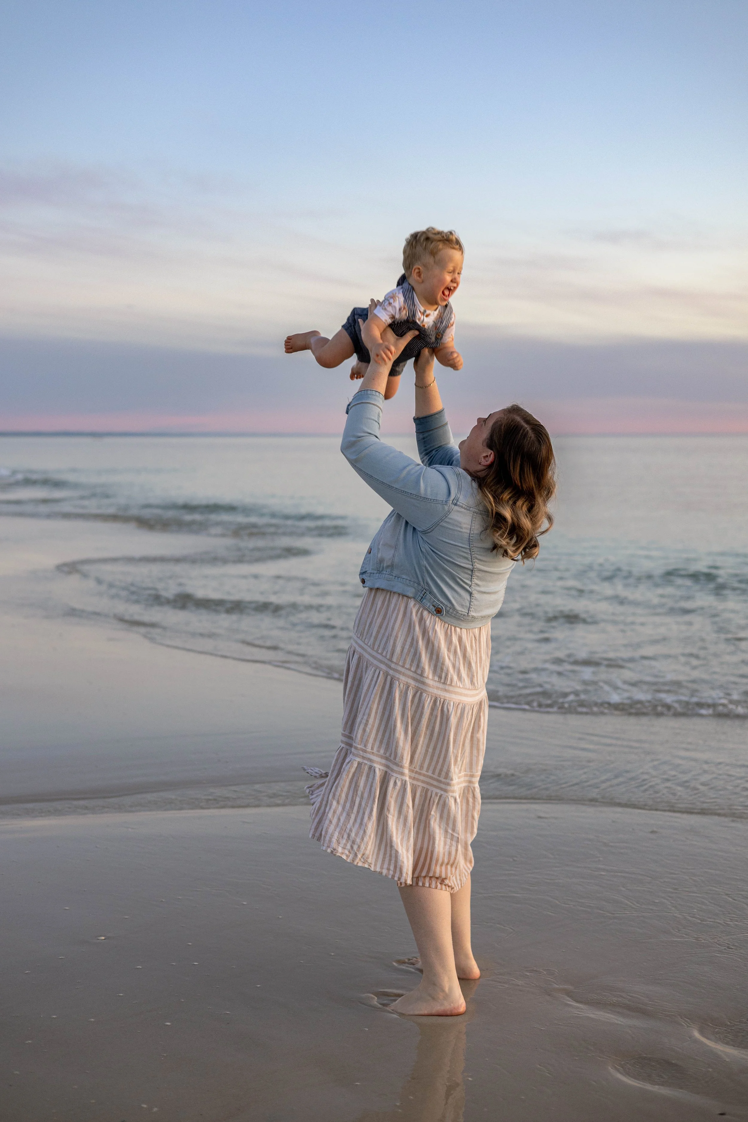 A woman on a beach holding a smiling child up in the air during sunset.