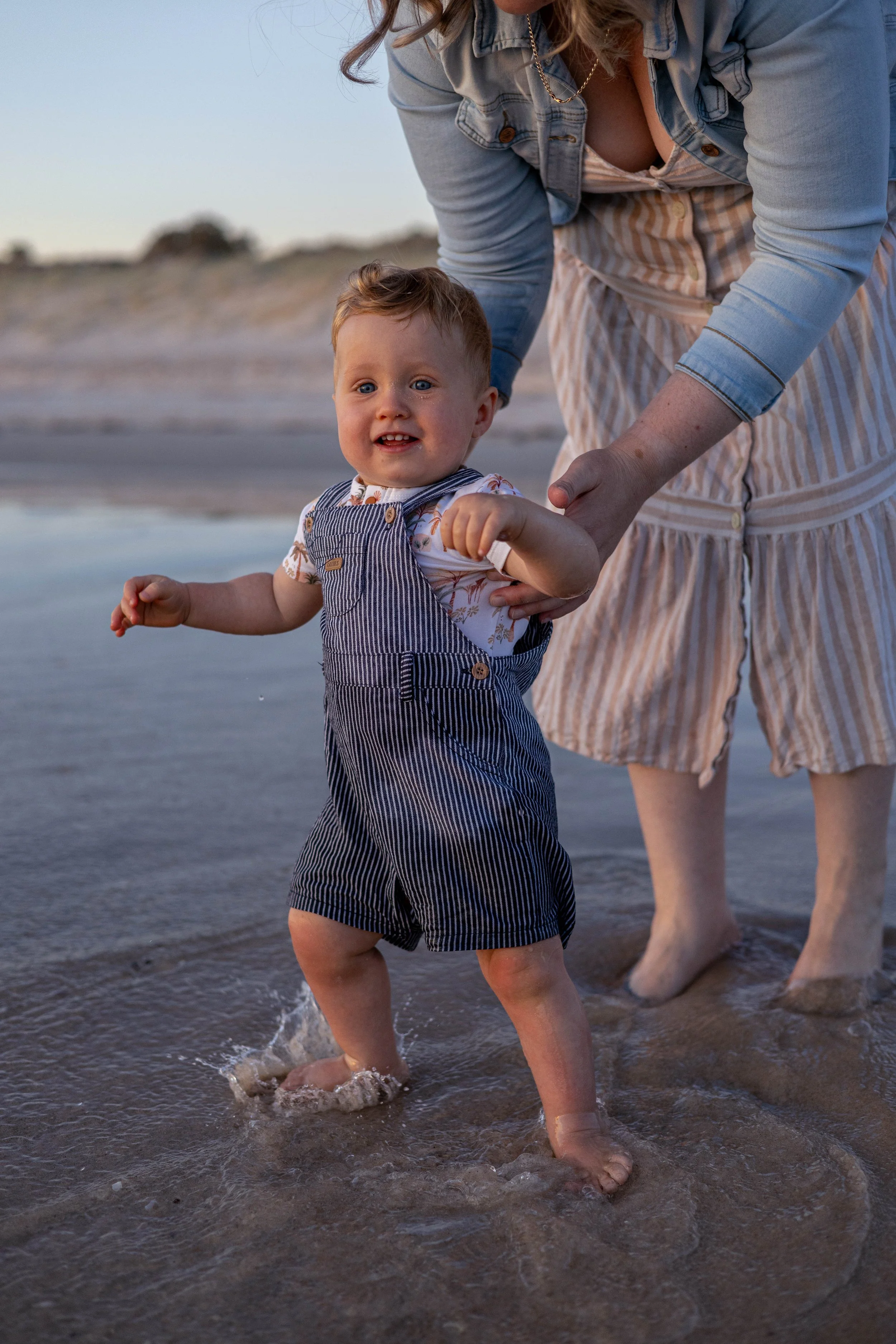 A toddler boy in striped overalls standing in shallow water at the beach, holding onto an adult woman's hand.