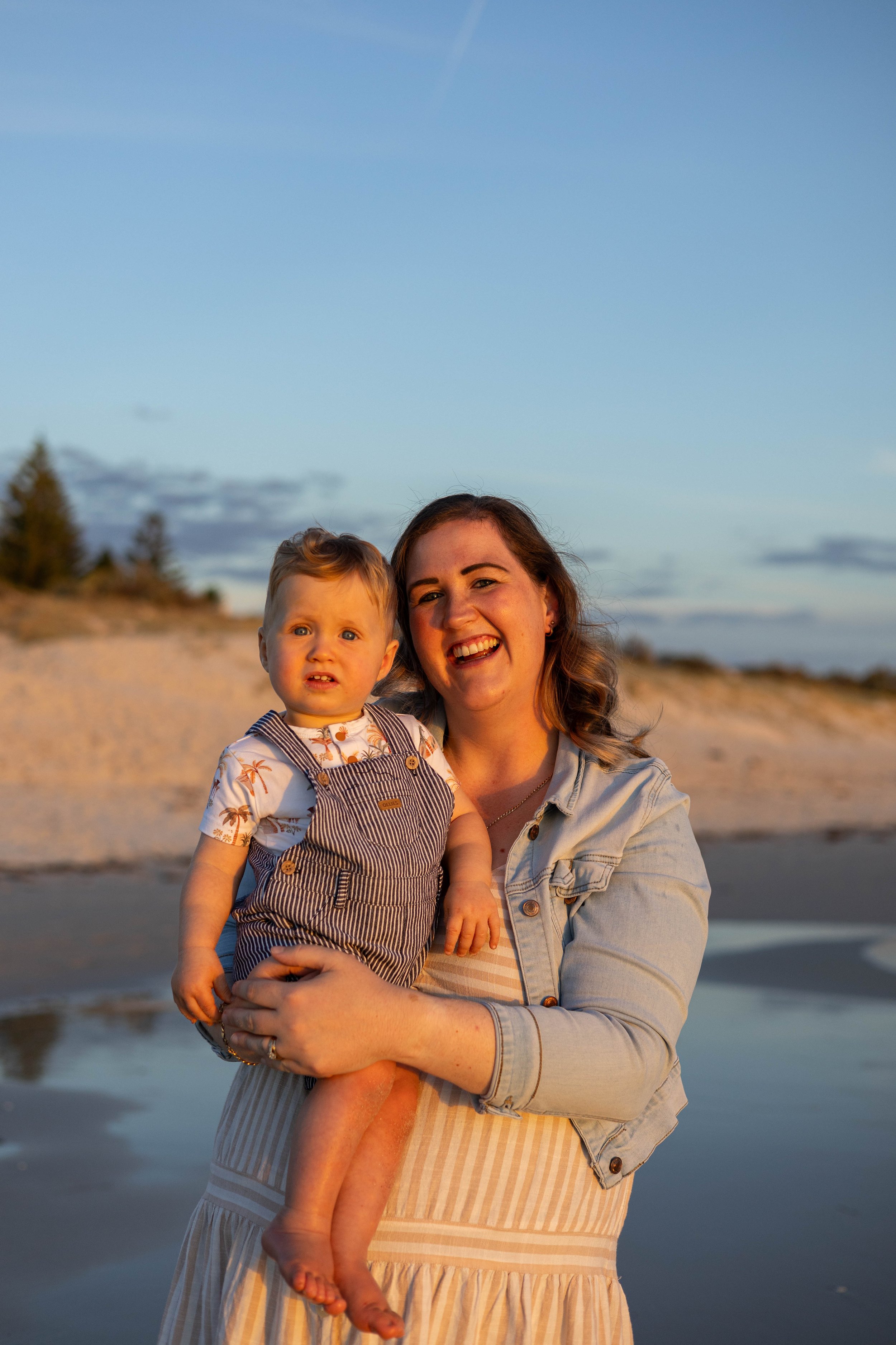 A woman holding a young boy on a beach during sunset, with dunes and trees in the background.
