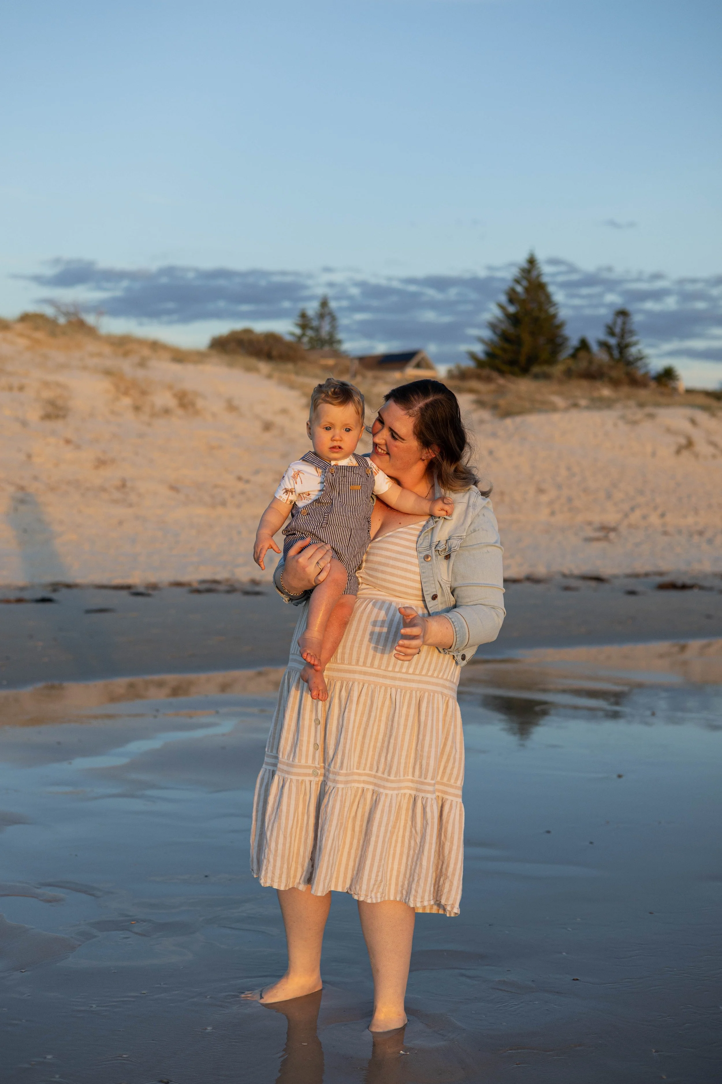 A woman holding a young child stands in shallow water at the beach during sunset, with sand dunes and trees in the background.