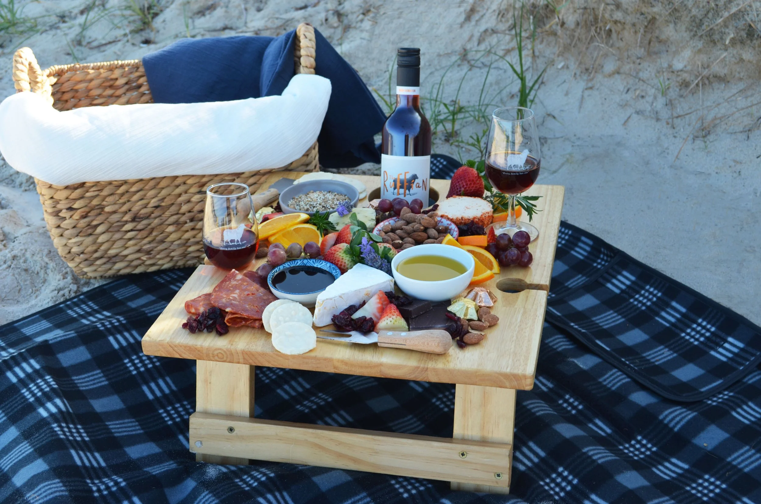 A picnic setup on a sandy beach with a low wooden table covered with a variety of cheeses, fruits, nuts, chocolates, and two glasses of red wine. There is a bottle of wine and a basket with a blue cloth and a white pillow, all set on a blue and black checkered blanket.
