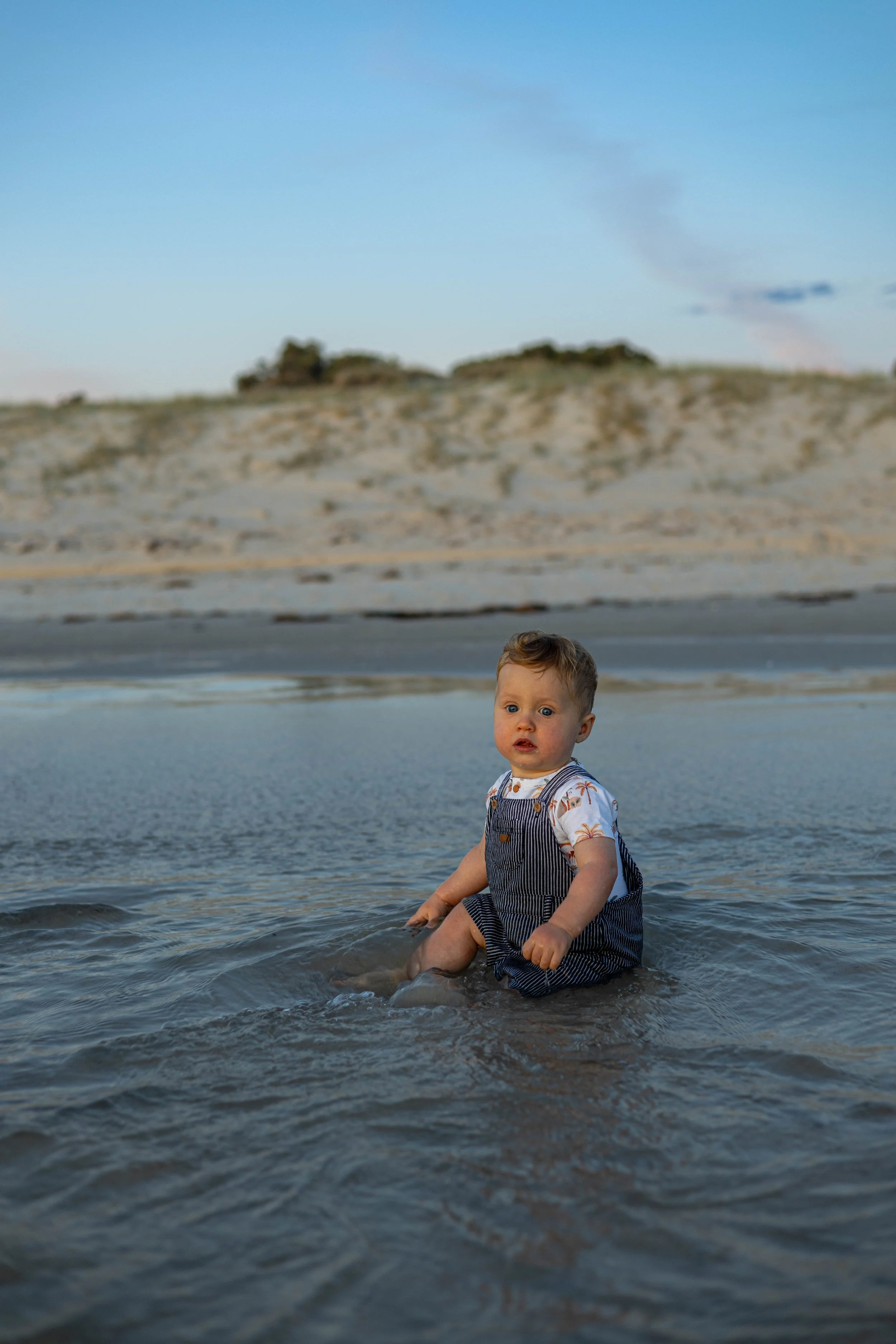 A baby sitting in shallow water at the beach, looking toward the camera with a curious expression.