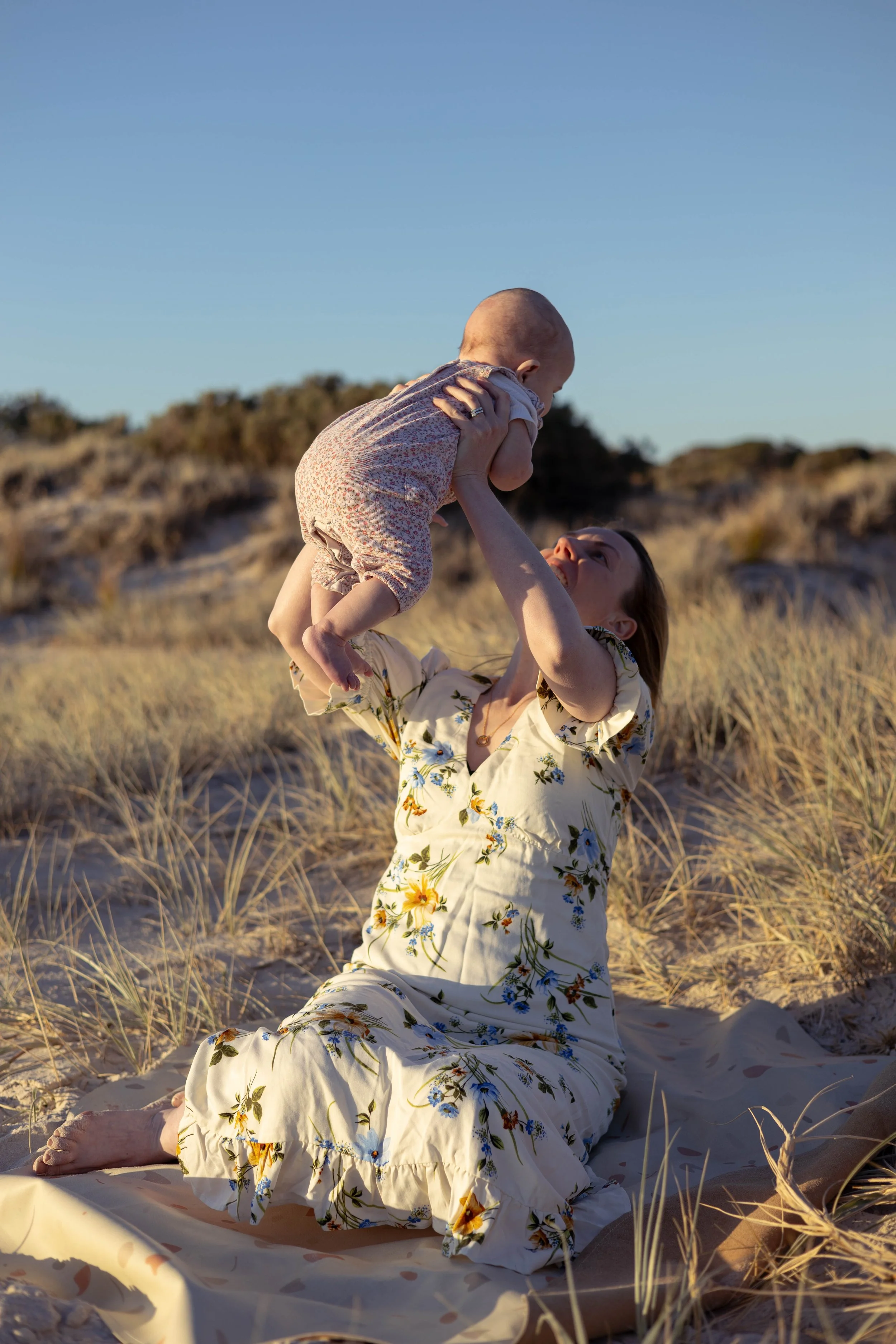 A woman in a floral dress sitting on sand in a grassy, beach-like area, lifting a baby in a patterned outfit above her with both hands, during sunset.