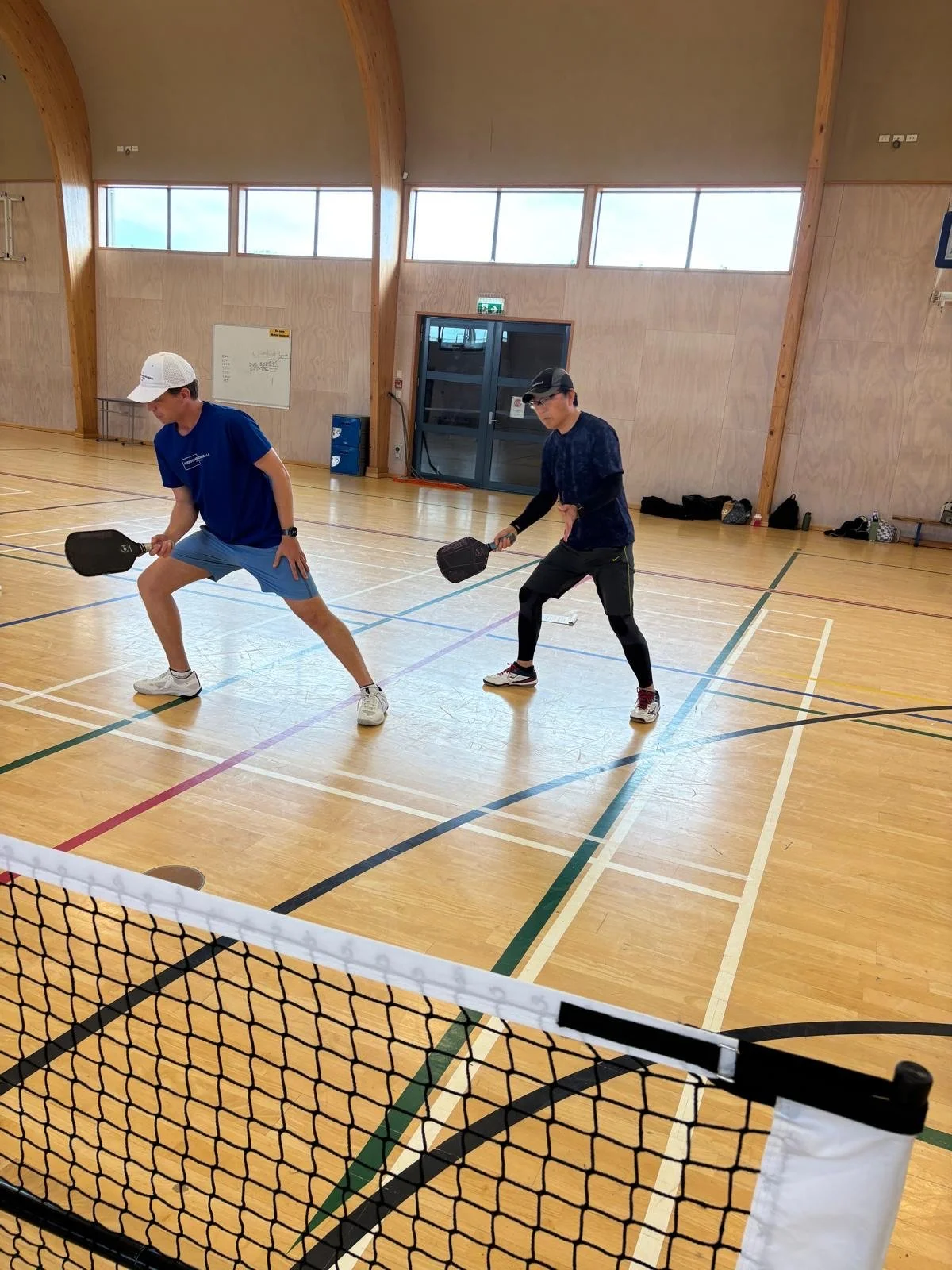 Coach Luke and a student playing pickleball indoors on a multi purpose court, holding paddles and ready to hit the ball.