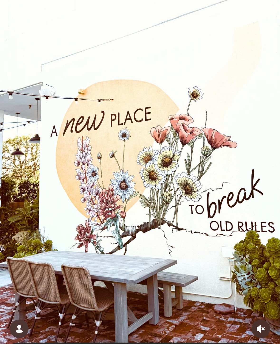 Decorative wall art with flowers and a message about breaking old rules, in a cozy outdoor seating area with a wooden table and chairs.