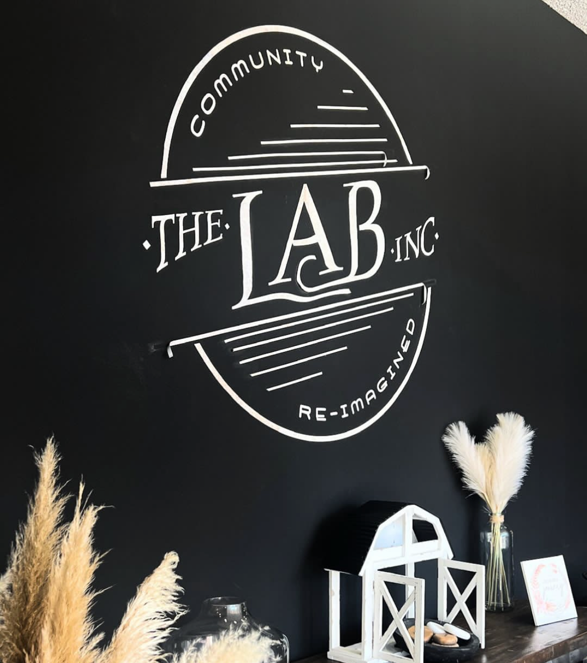 Decorative wall sign with the text 'The Lab Inc.' and the slogans 'Community' and 'Re-imagined' in a stylish design, surrounded by dried pampas grass and small decorative items on a table.