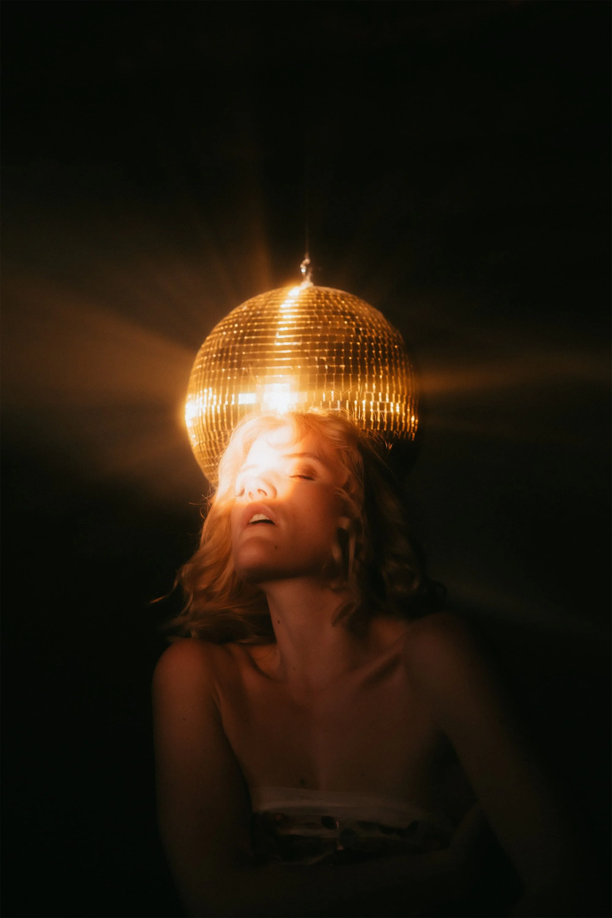 A woman with closed eyes is illuminated by a glowing disco ball behind her head, creating a radiant light effect.