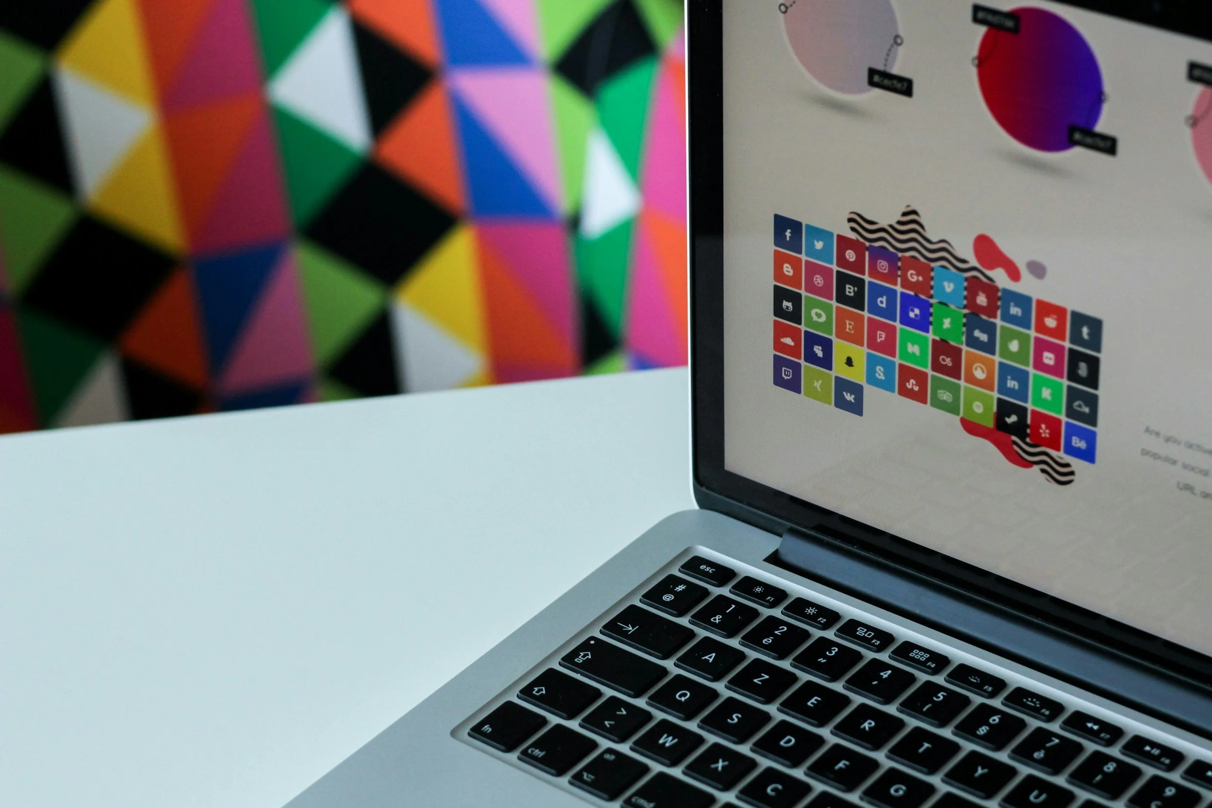 Close-up of a laptop showing a colorful grid of social media icons on the screen, placed on a white surface with a blurred multicolored geometric patterned background.