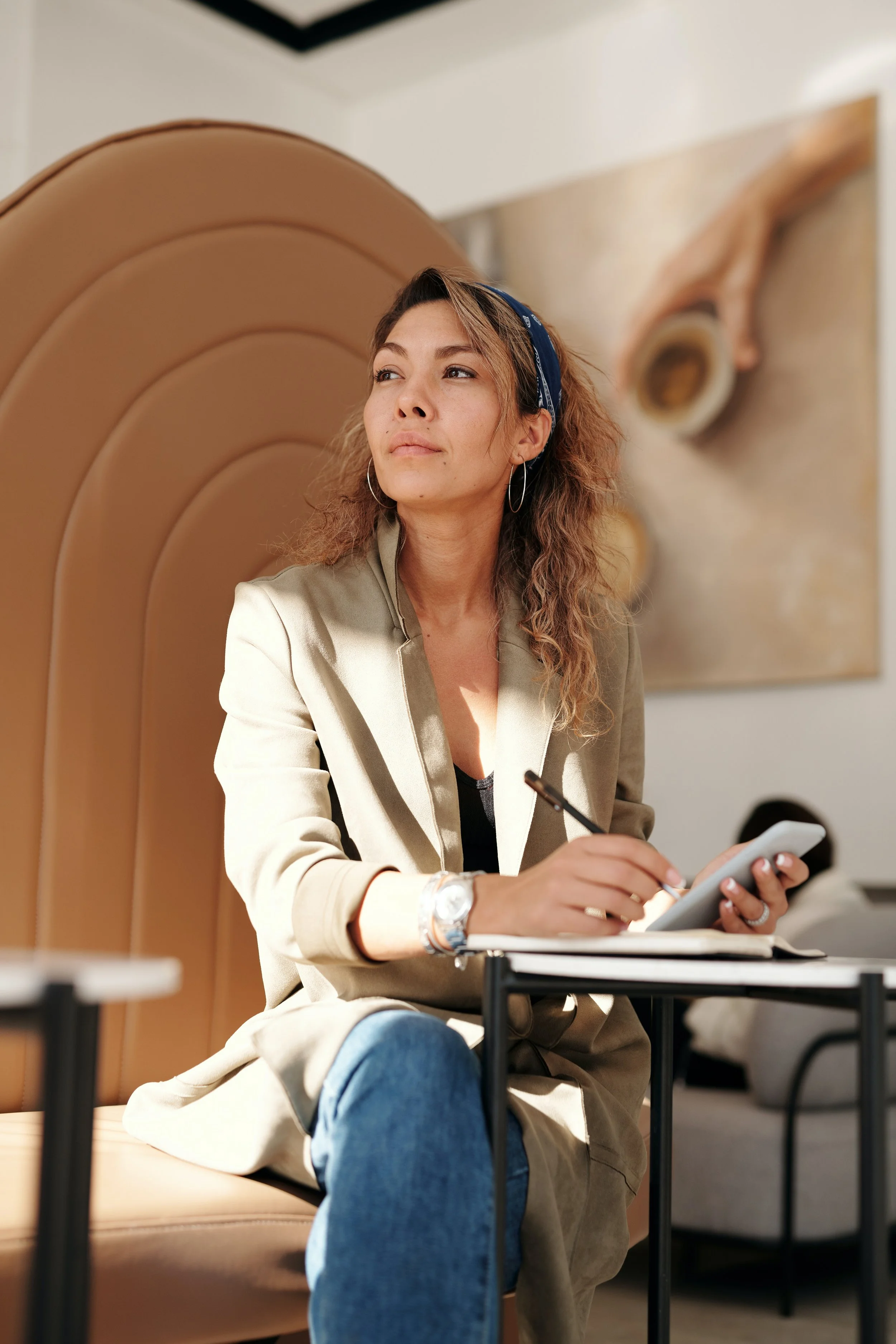 A woman with curly hair, wearing a beige jacket, blue bandana, and hoop earrings, sitting on a beige chair, looking thoughtfully to the side while holding a phone and pen in her hands, with a large painting of an animal's eye in the background.