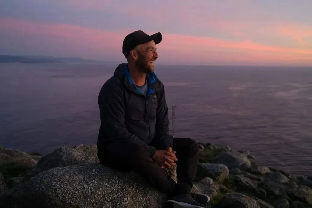 A man wearing a black cap and jacket sitting on rocks by the water during sunset, smiling and looking to the left.