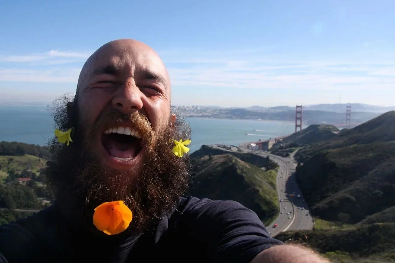 Man with a beard and flowers in his hair, laughing in front of a scenic view of the Golden Gate Bridge and San Francisco Bay.