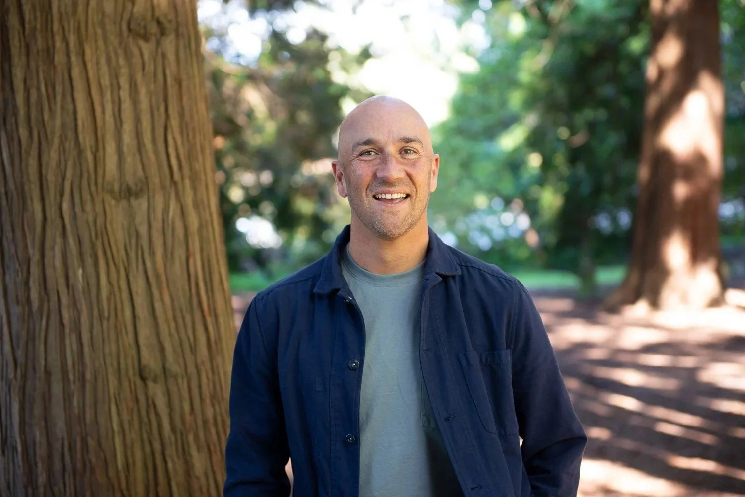 A man with a shaved head and beard smiling outdoors in a park with large trees and sunlight.