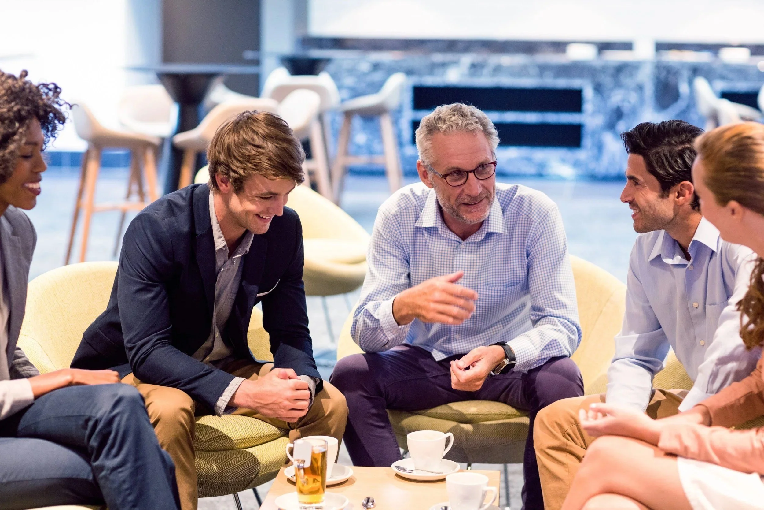 A group of diverse people sitting together on a yellow couch, engaged in conversation, smiling, in a modern office or lounge space with coffee cups and a table in front of them.