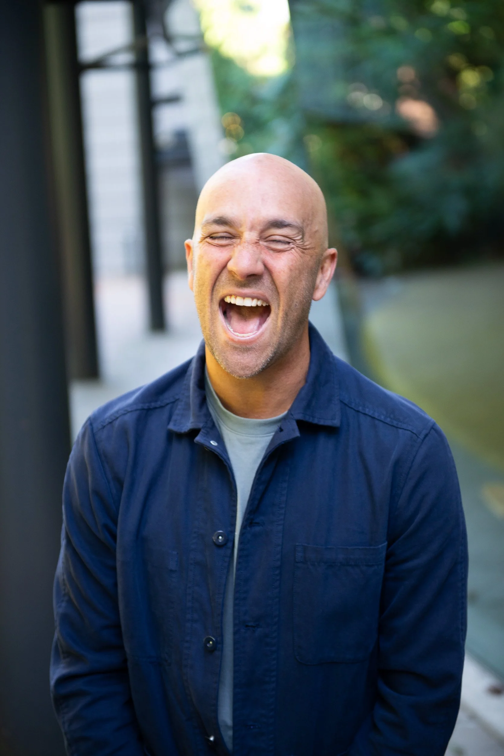 A bald man in a blue denim shirt laughing outdoors during the daytime.
