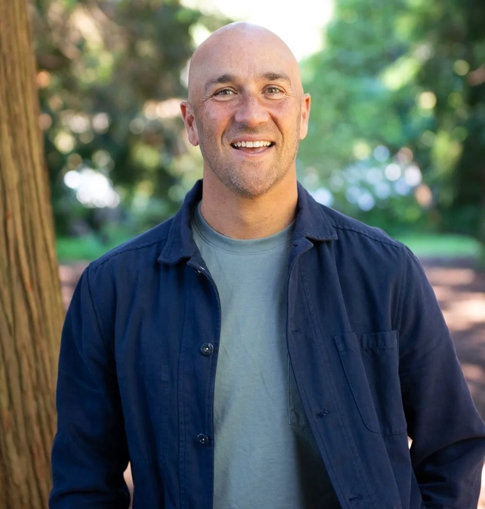 A smiling bald man in a light gray t-shirt and navy button-up shirt standing outdoors in a green park.